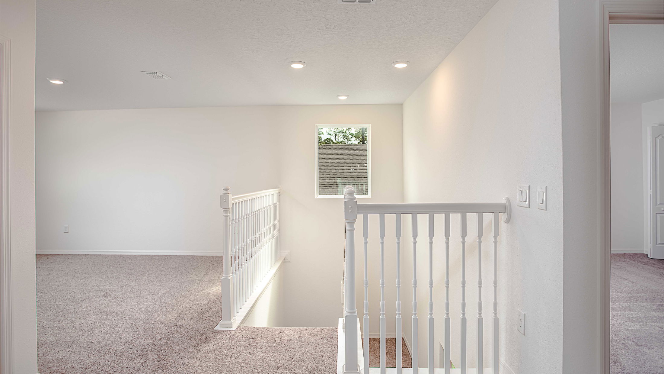 Bright, modern upper floor hallway with carpeted flooring, recessed lighting, and a white wooden stair railing next to a window.