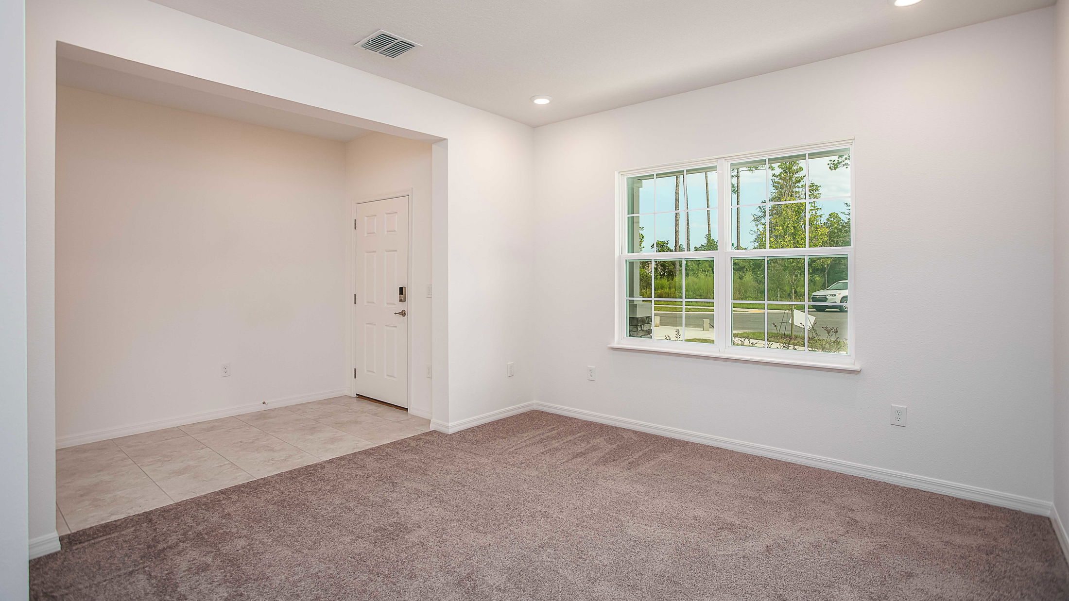 Empty room with plush carpet, white walls, and a large window offering natural light, adjacent to a tiled entrance.