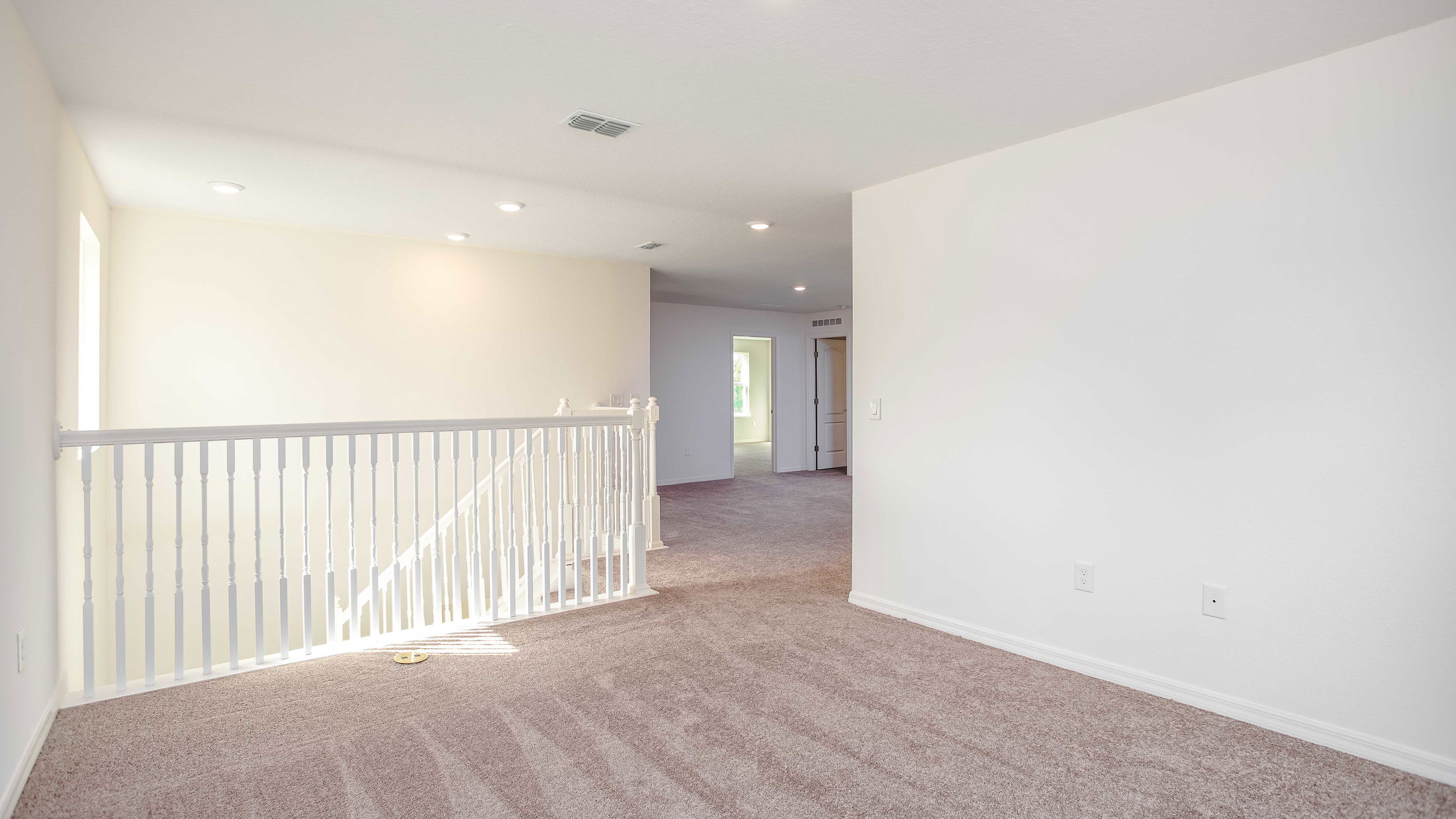 Spacious carpeted upstairs hallway with white railings and recessed lighting, leading to adjacent rooms in a modern home.