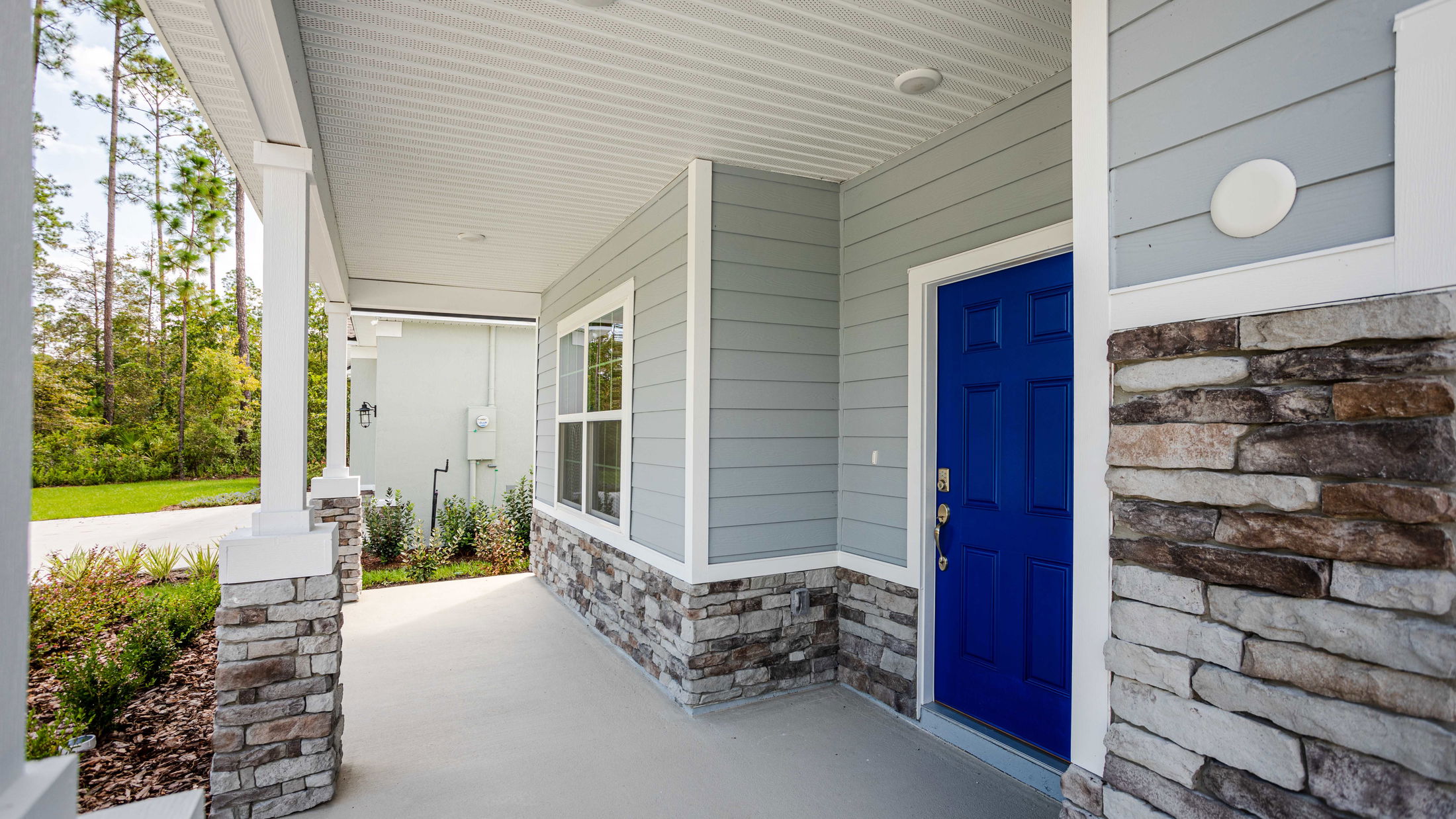 Front porch of a modern house with gray siding, stone accents, and a vibrant blue door surrounded by green landscaping.