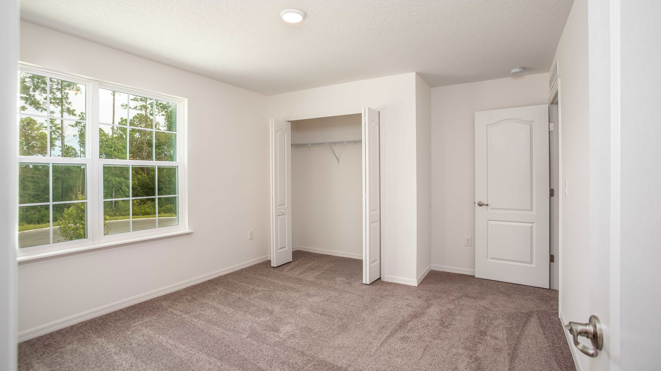 Empty white-walled bedroom with plush carpet, open closet, and large window overlooking greenery.