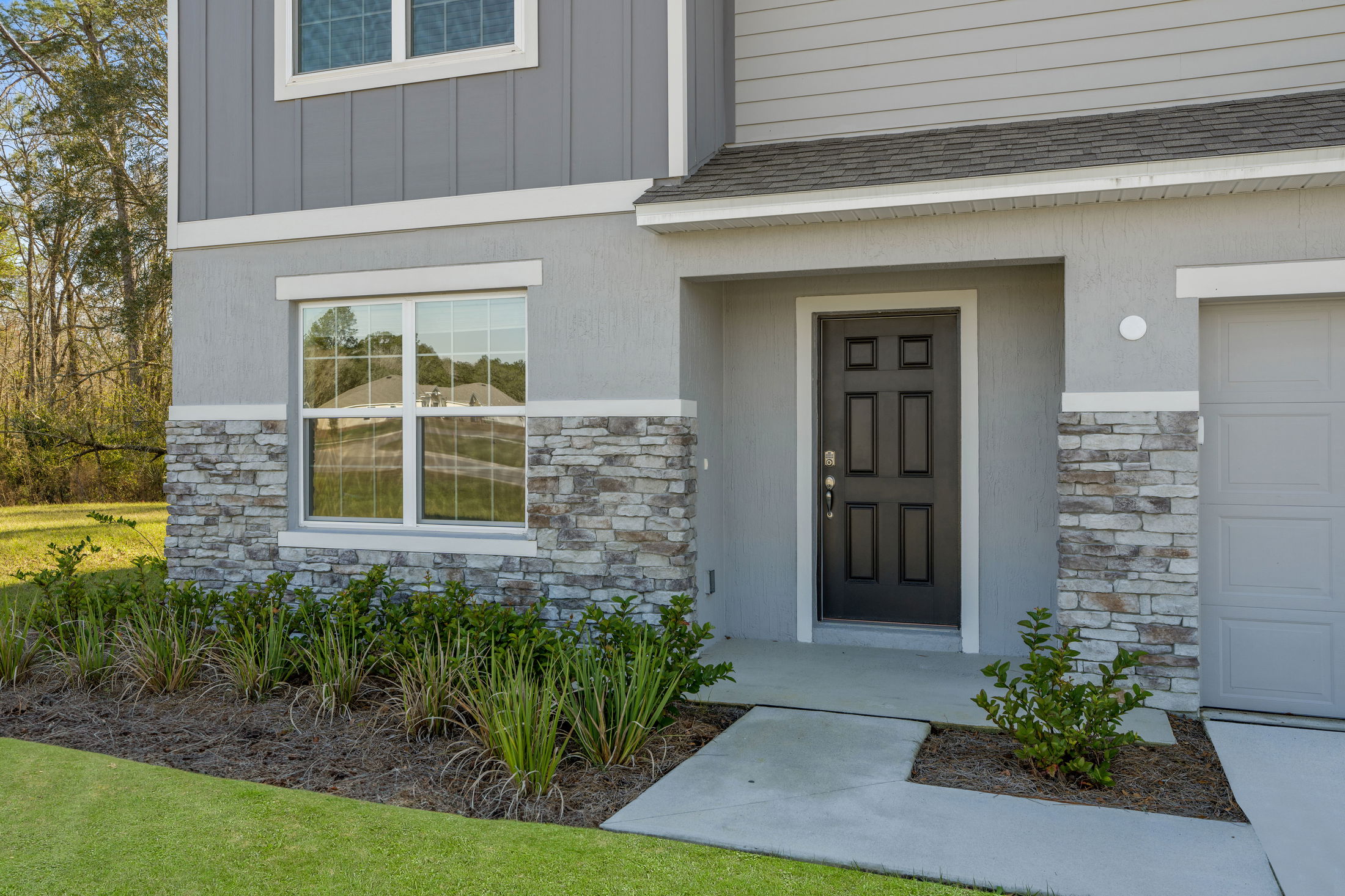 Close-up of a modern house exterior featuring a black front door, stone and siding facade, and a landscaped garden with green plants.