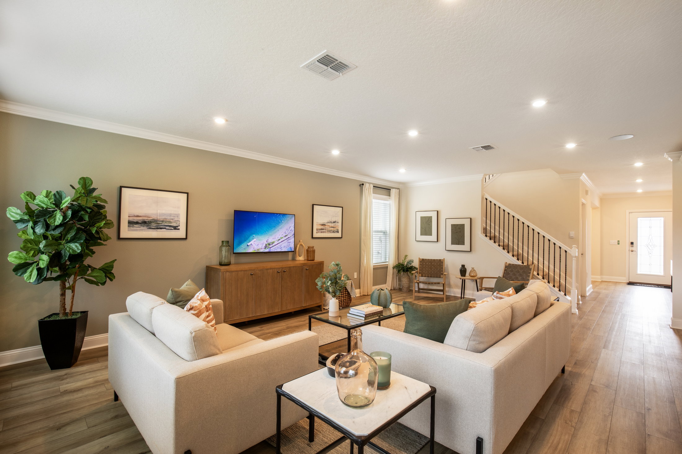Bright living room featuring a beige sofa set, a wooden entertainment unit with a TV, and decorative plants.
