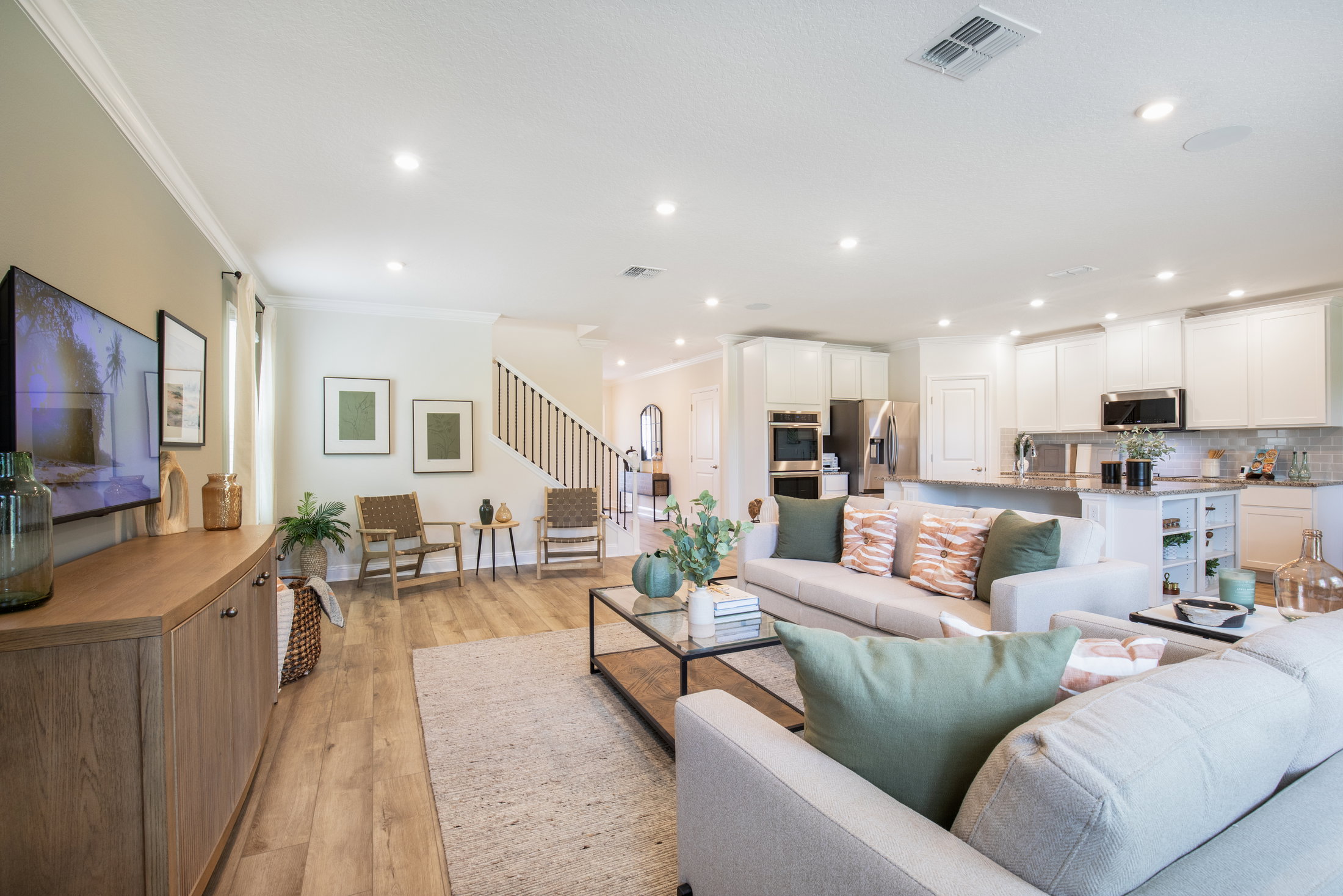 A modern living room and kitchen area featuring light-colored furniture, a coffee table with a plant, and a large TV on a wooden console.
