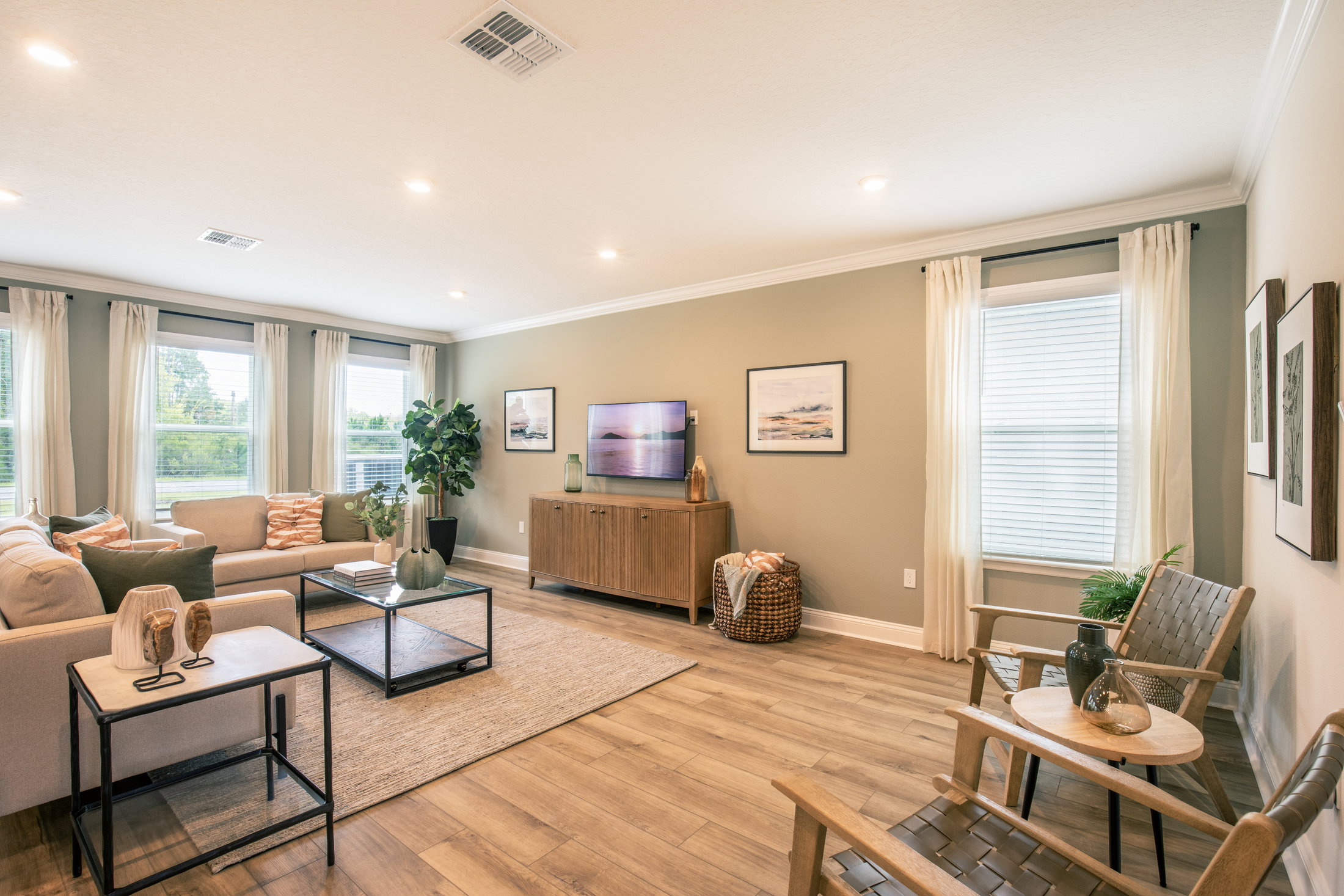 A modern living room featuring a sofa, coffee table, two chairs, decorative plants, and large windows with curtains.
