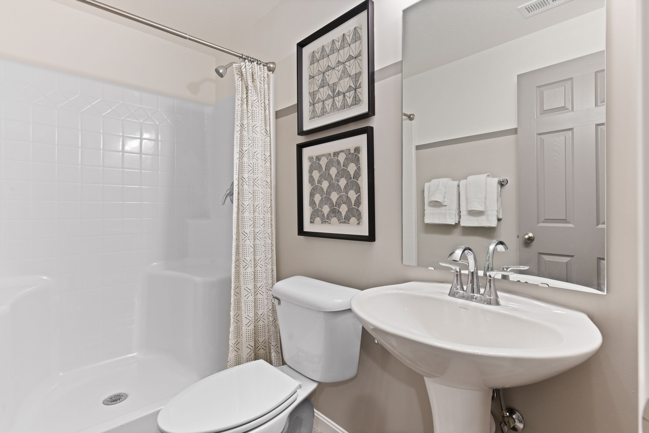 A modern bathroom featuring a white pedestal sink, a toilet, a shower with patterned curtain, and geometric wall art.