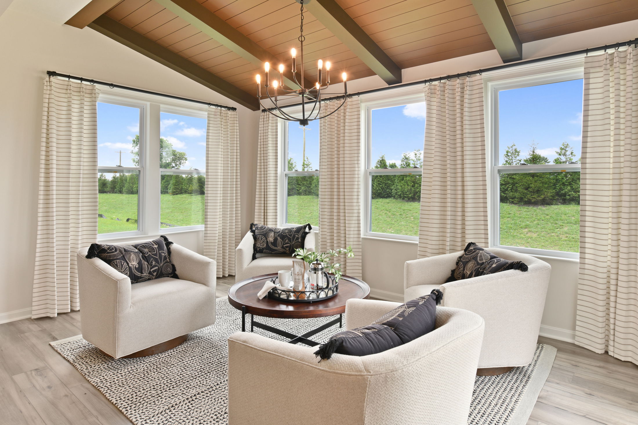 Cozy modern living room with beige armchairs, a round wooden coffee table, a chandelier, and large windows overlooking a green lawn.