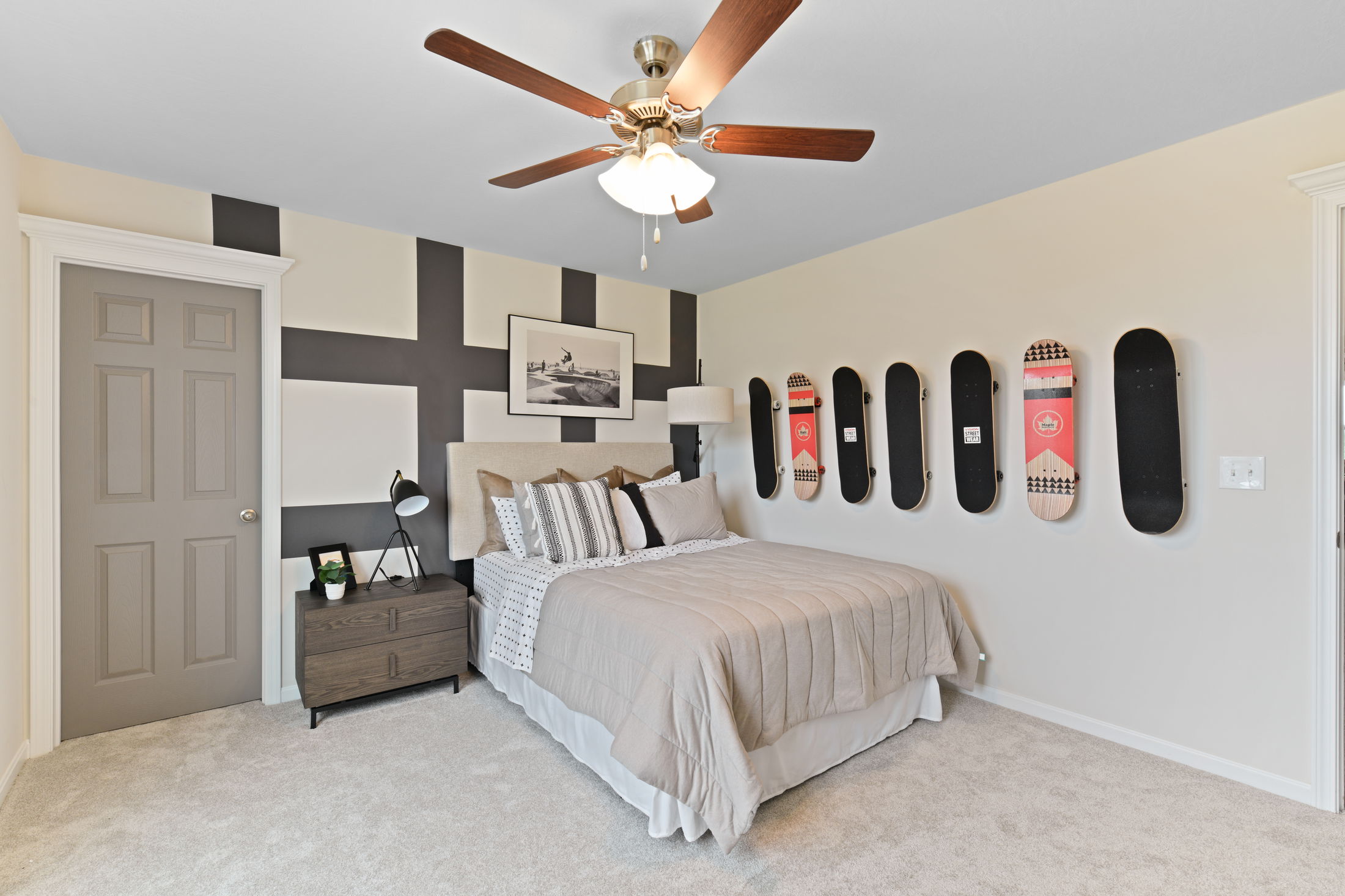 Modern bedroom with a beige bed, skateboard decor on the wall, geometric accent wall, ceiling fan, and wooden nightstand.