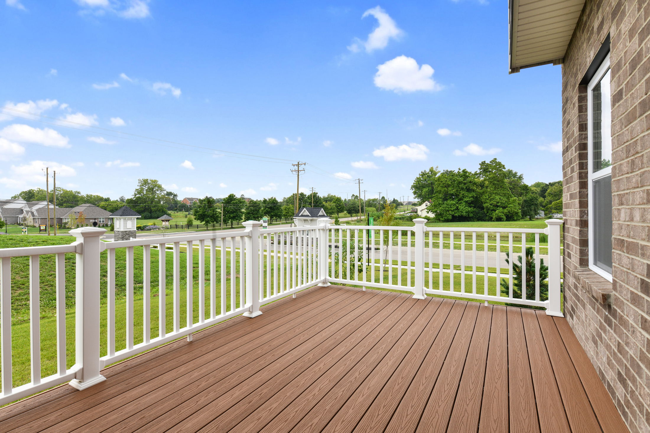 A spacious outdoor deck with white railing overlooks a lush green neighborhood landscape under a clear blue sky.