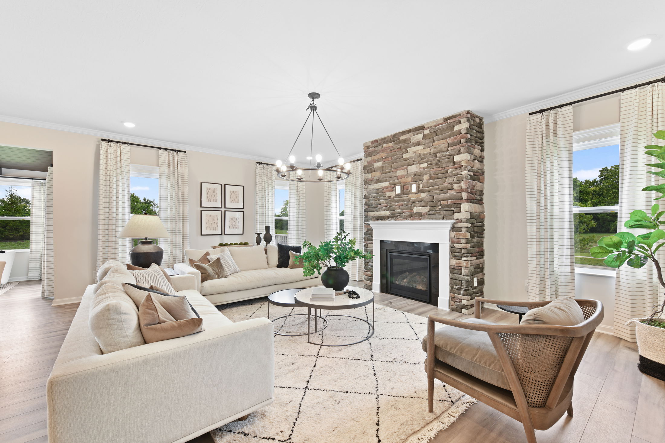 Modern living room with a stone fireplace, beige sofas, wooden chair, and large windows framed by striped curtains, accented by a round chandelier and potted greenery.