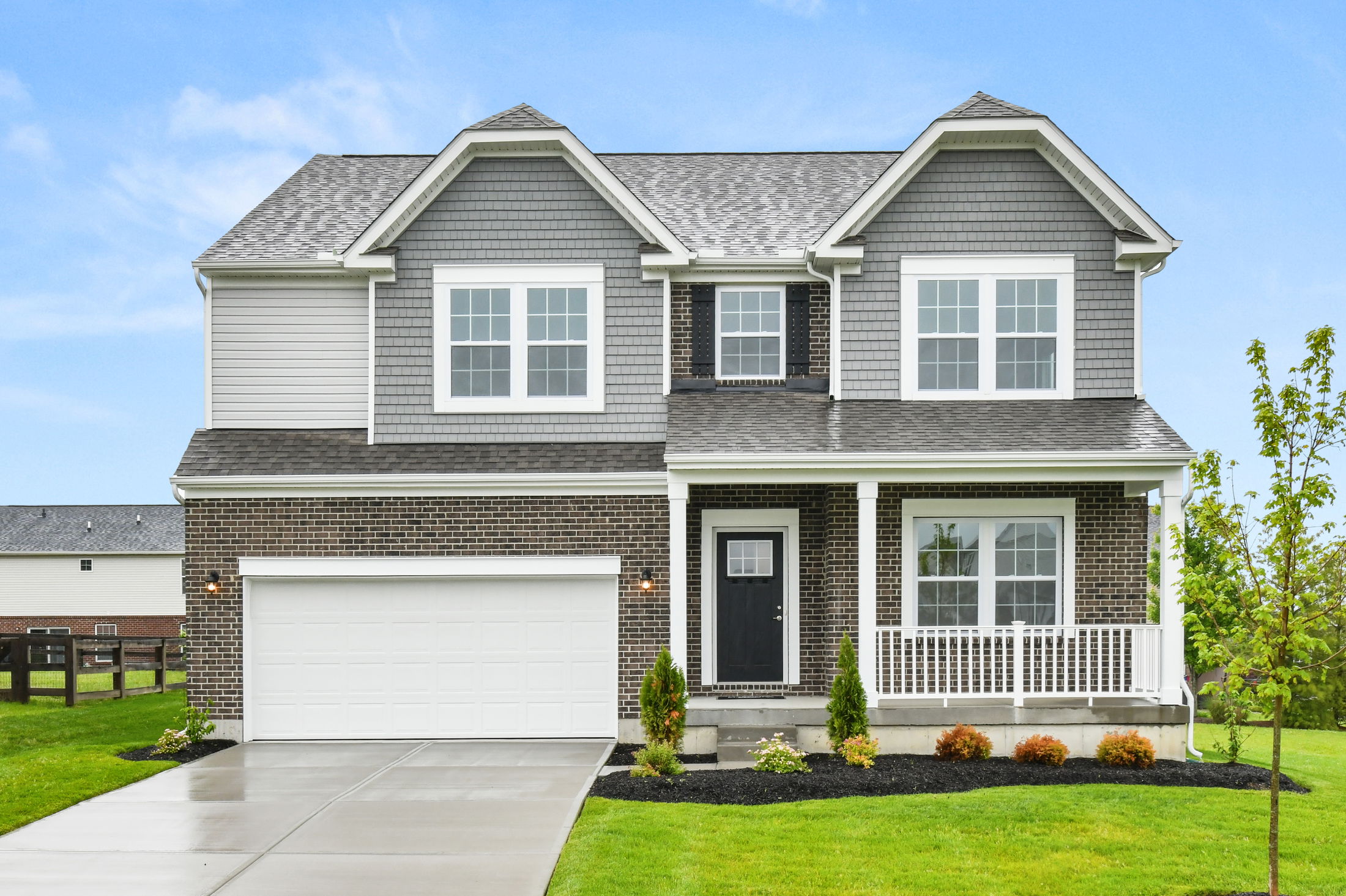 Two-story suburban house with a gray facade, front porch, and attached garage on a sunny day.