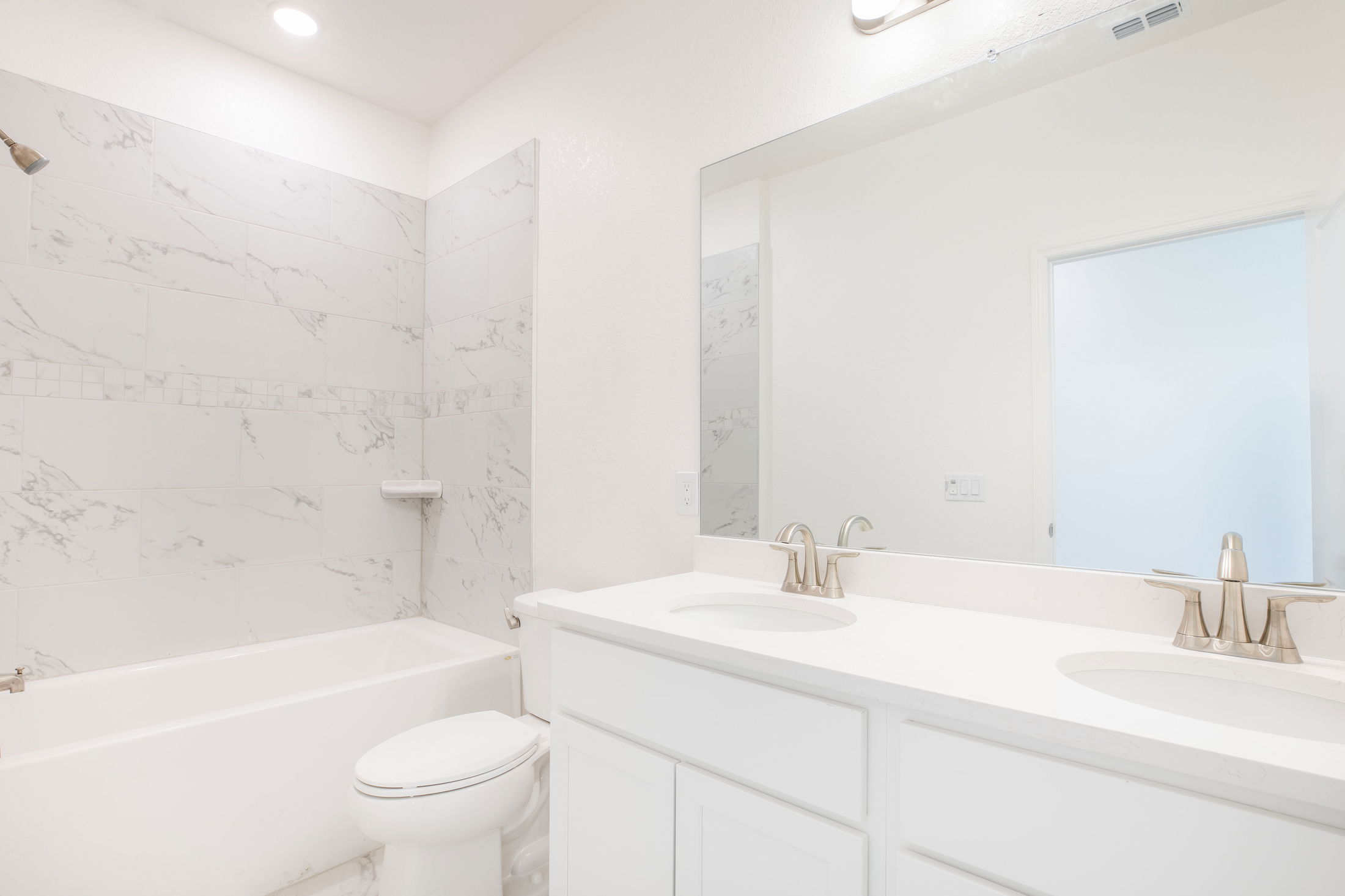 Modern white bathroom with double sinks, a spacious mirror, and a bathtub with marble tile walls.