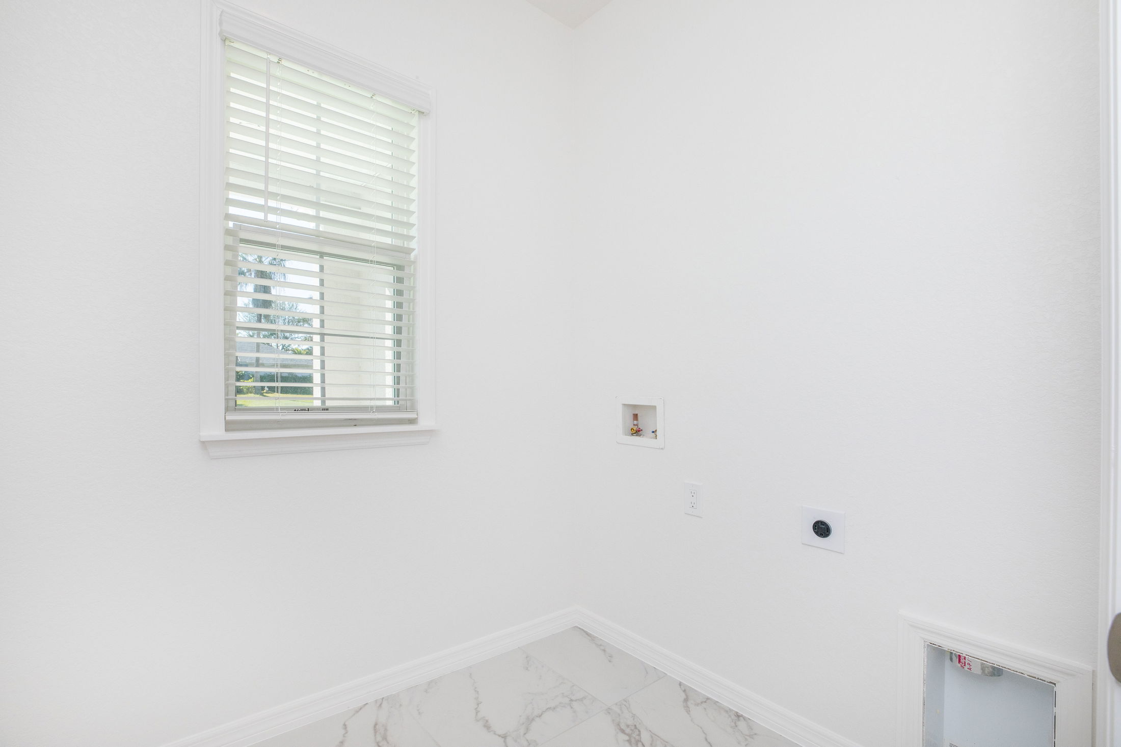 Empty laundry room with white walls, a small window with blinds, and hookups for appliances on a marble tile floor.