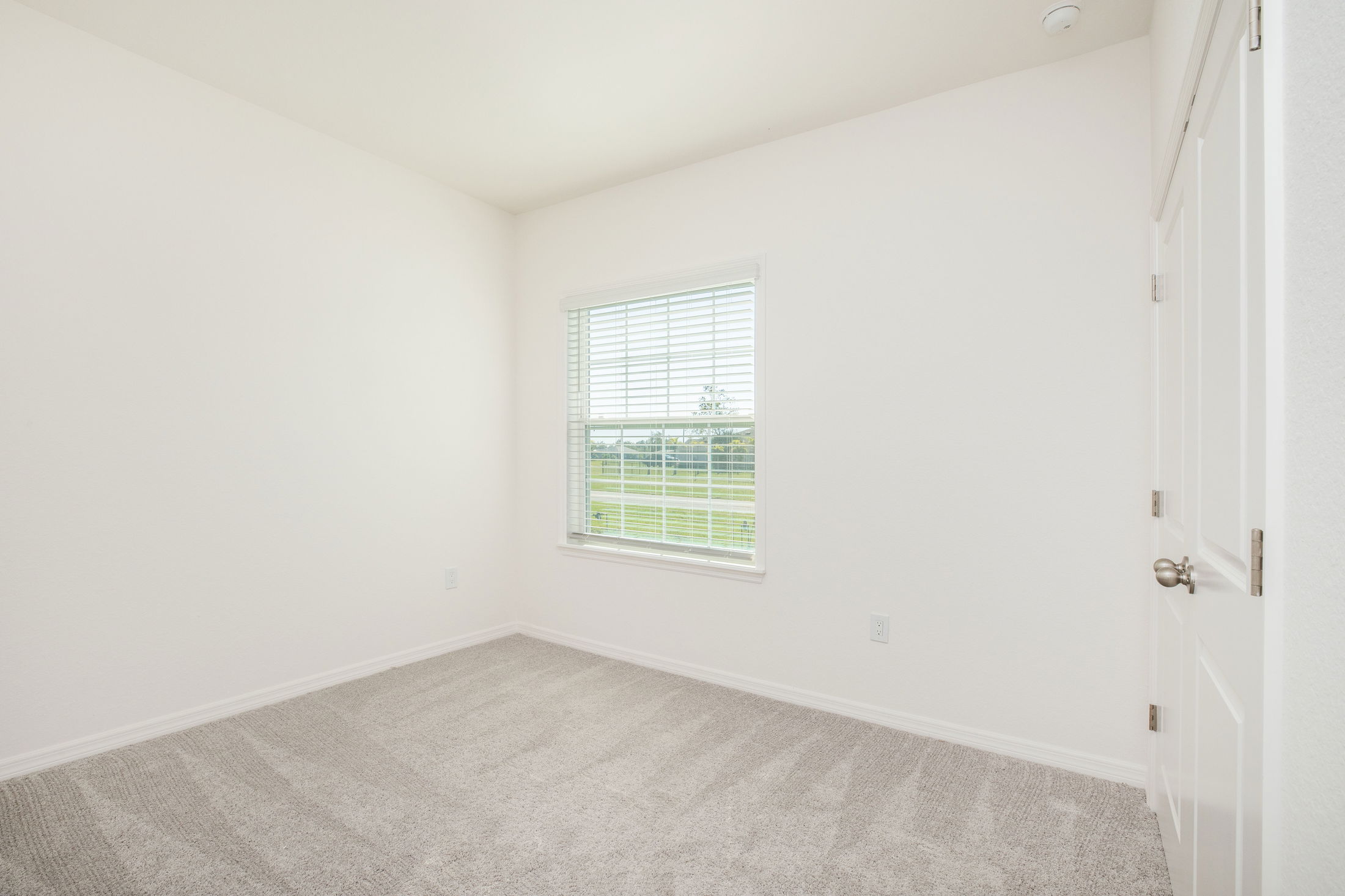 Empty, bright room with a beige carpet, white walls, and a window with blinds.
