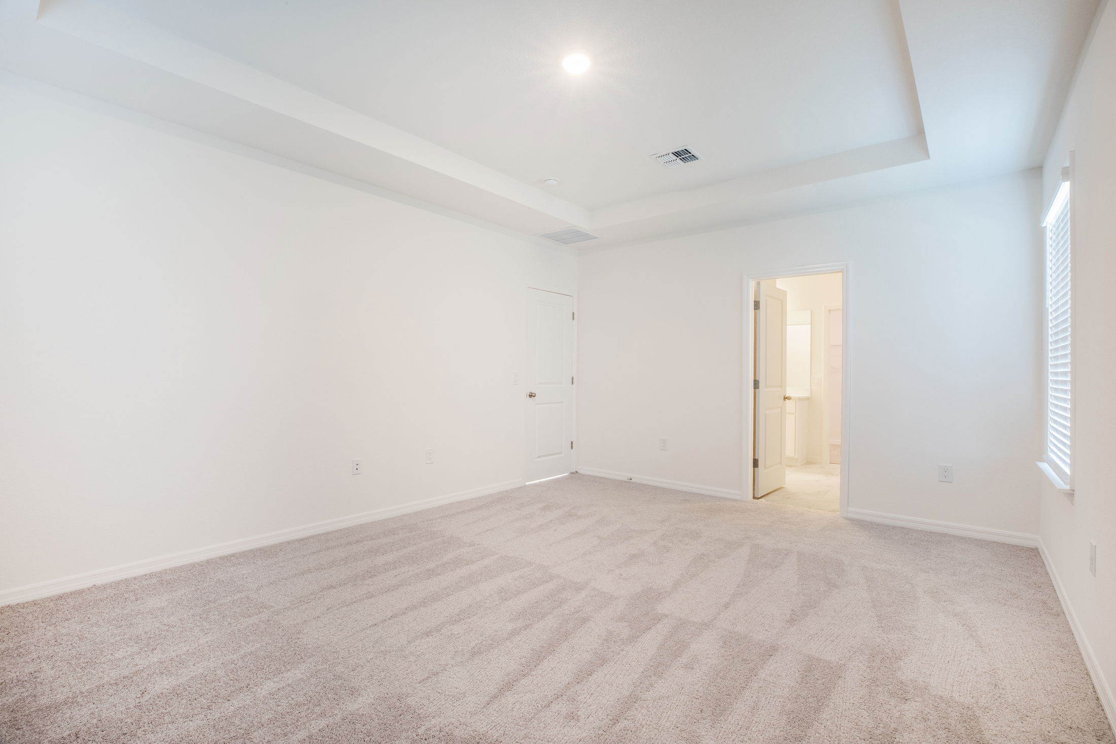 Empty, bright carpeted room with a white ceiling, recessed lighting, and an open door leading to a bathroom.