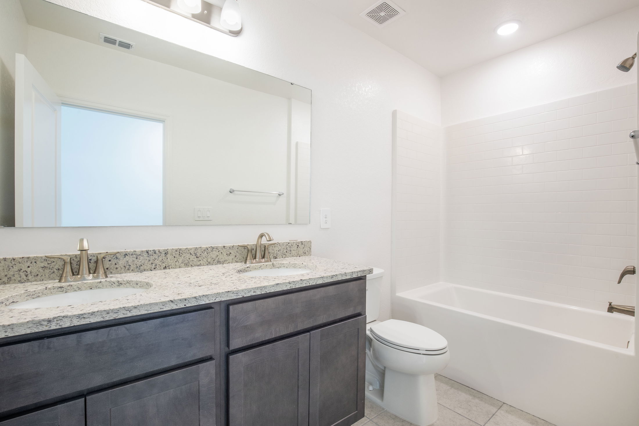 Modern bathroom with granite countertops, double sinks, a large mirror, and a white bathtub with tile surround.