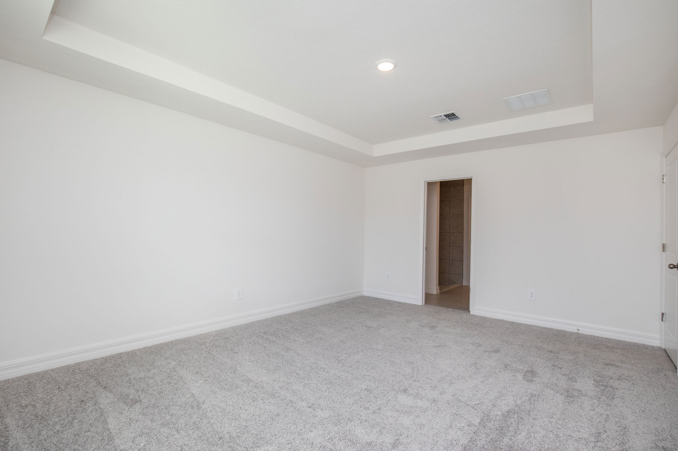 Spacious empty room with white walls, gray carpet, and a recessed ceiling leading to a tiled bathroom.