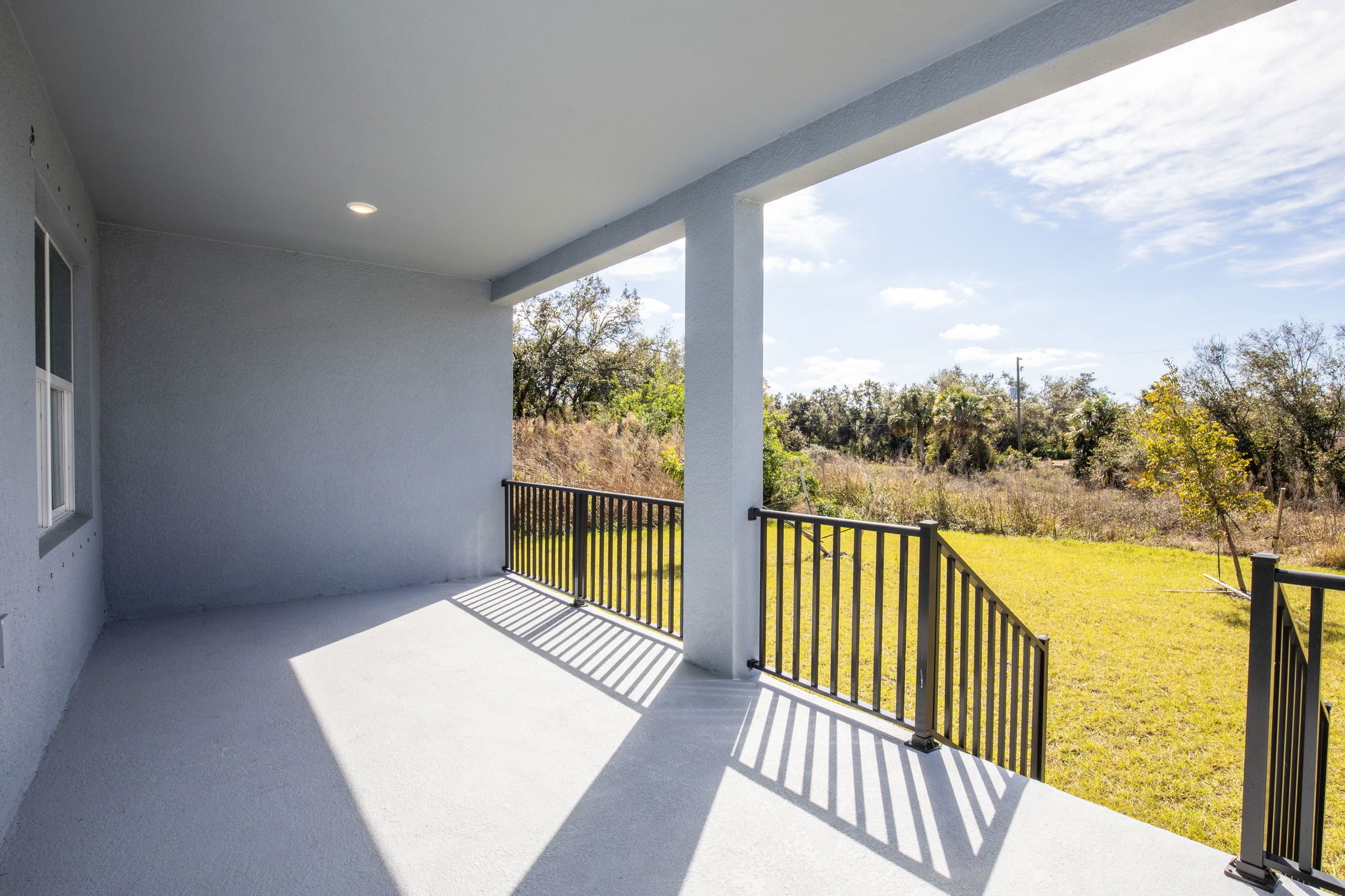 Covered patio with black railing overlooking a sunny yard and natural landscape in the background.