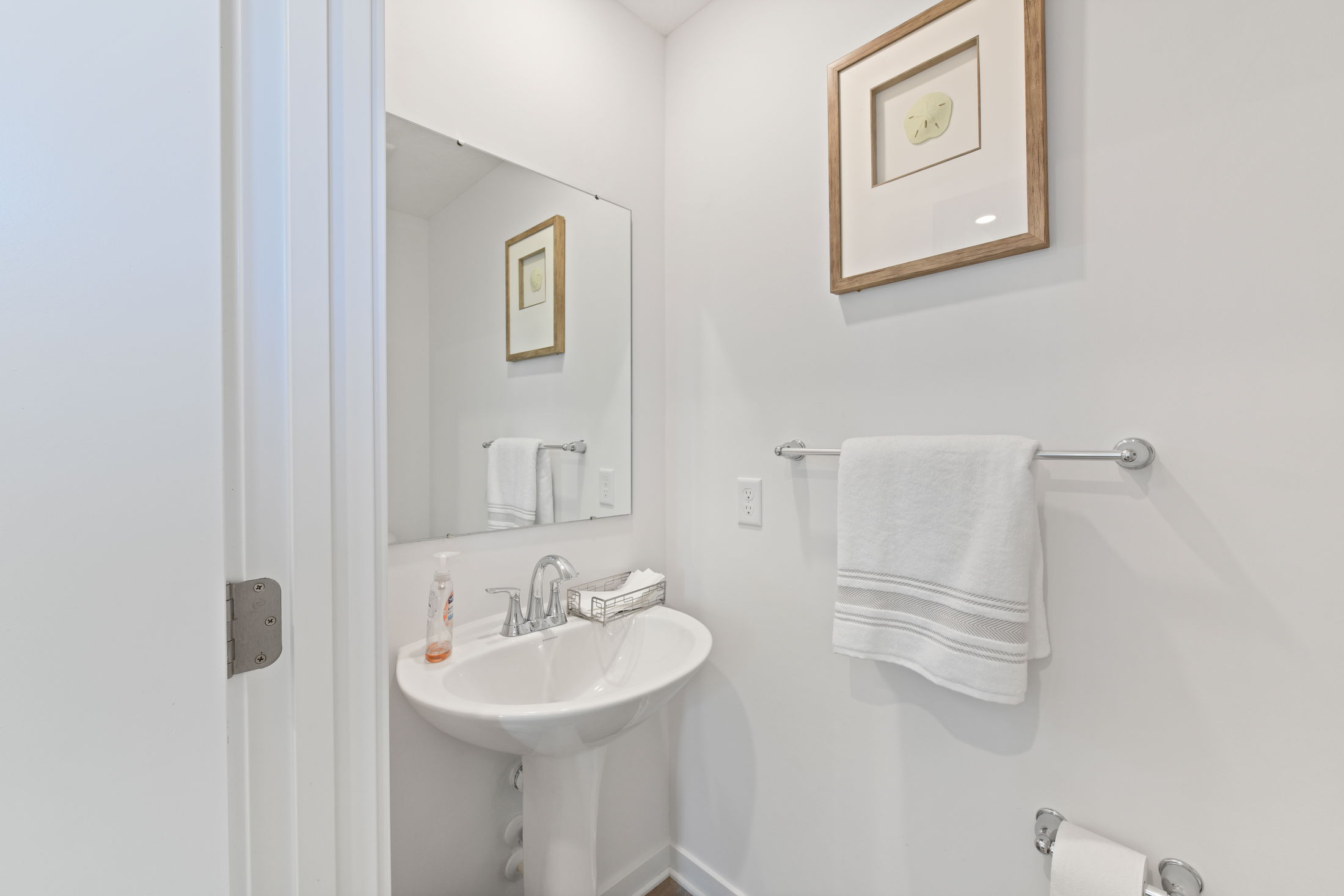 Minimalist white bathroom with pedestal sink, large mirror, and wall art over towel rack.