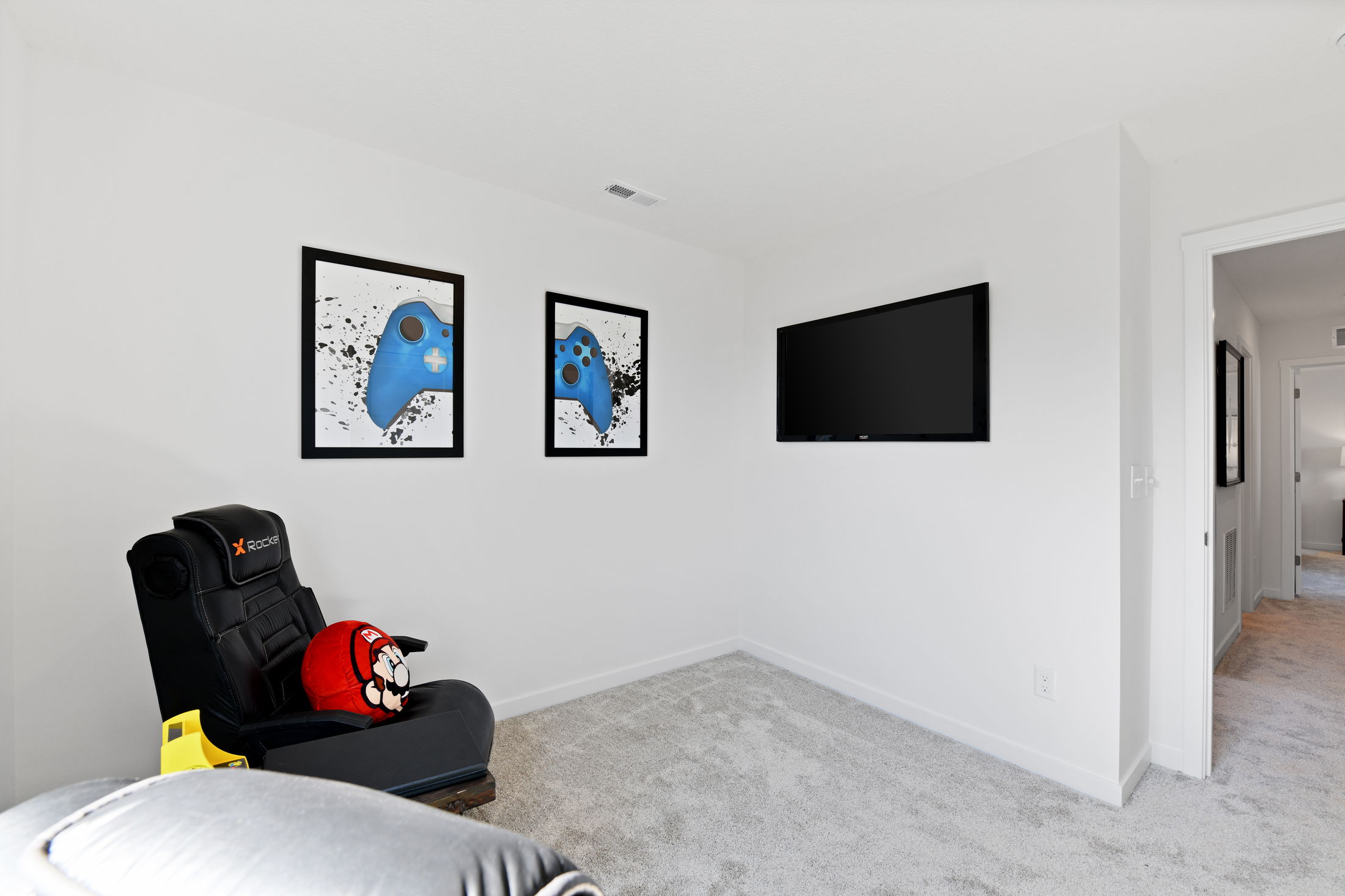 Gaming room with X Rocker chair, wall-mounted TV, framed blue gaming controller art, and a Mario-themed plush toy.