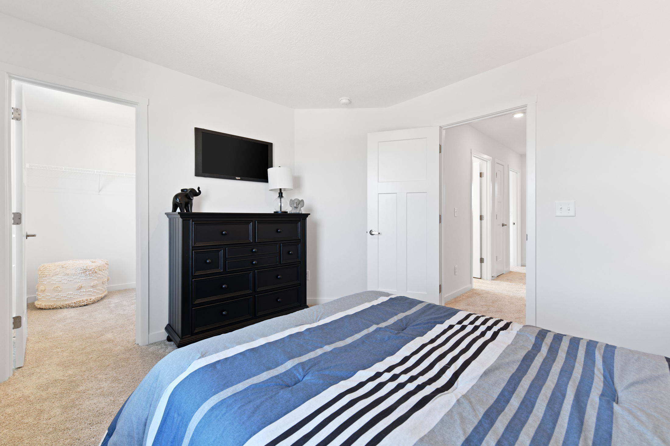 A modern bedroom with white walls, a striped blue bedspread, black dresser, TV, and open doors to a walk-in closet and hallway.