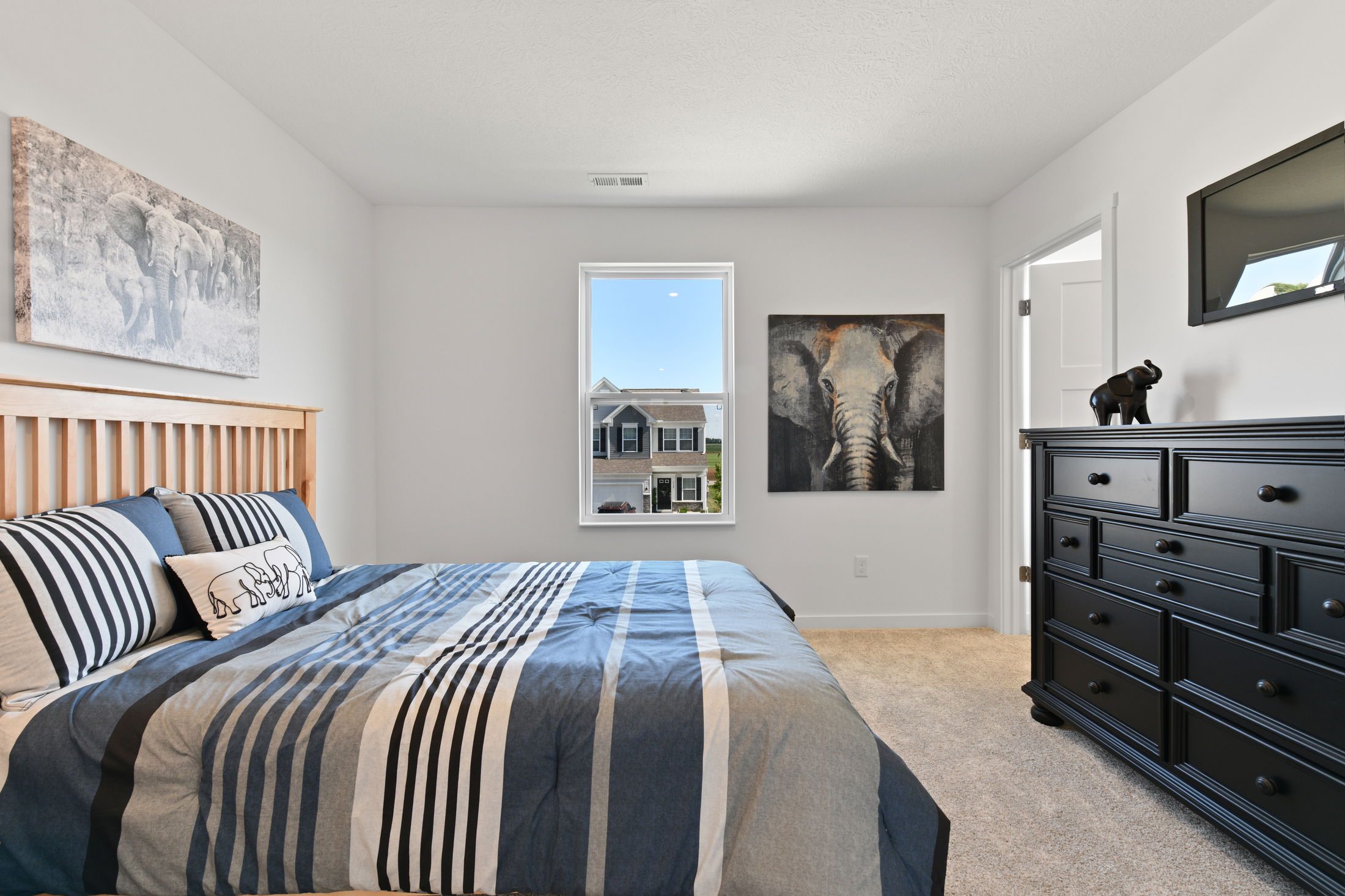 Cozy bedroom with striped bedspread, elephant-themed decor, black dresser, and a window view of neighboring house.