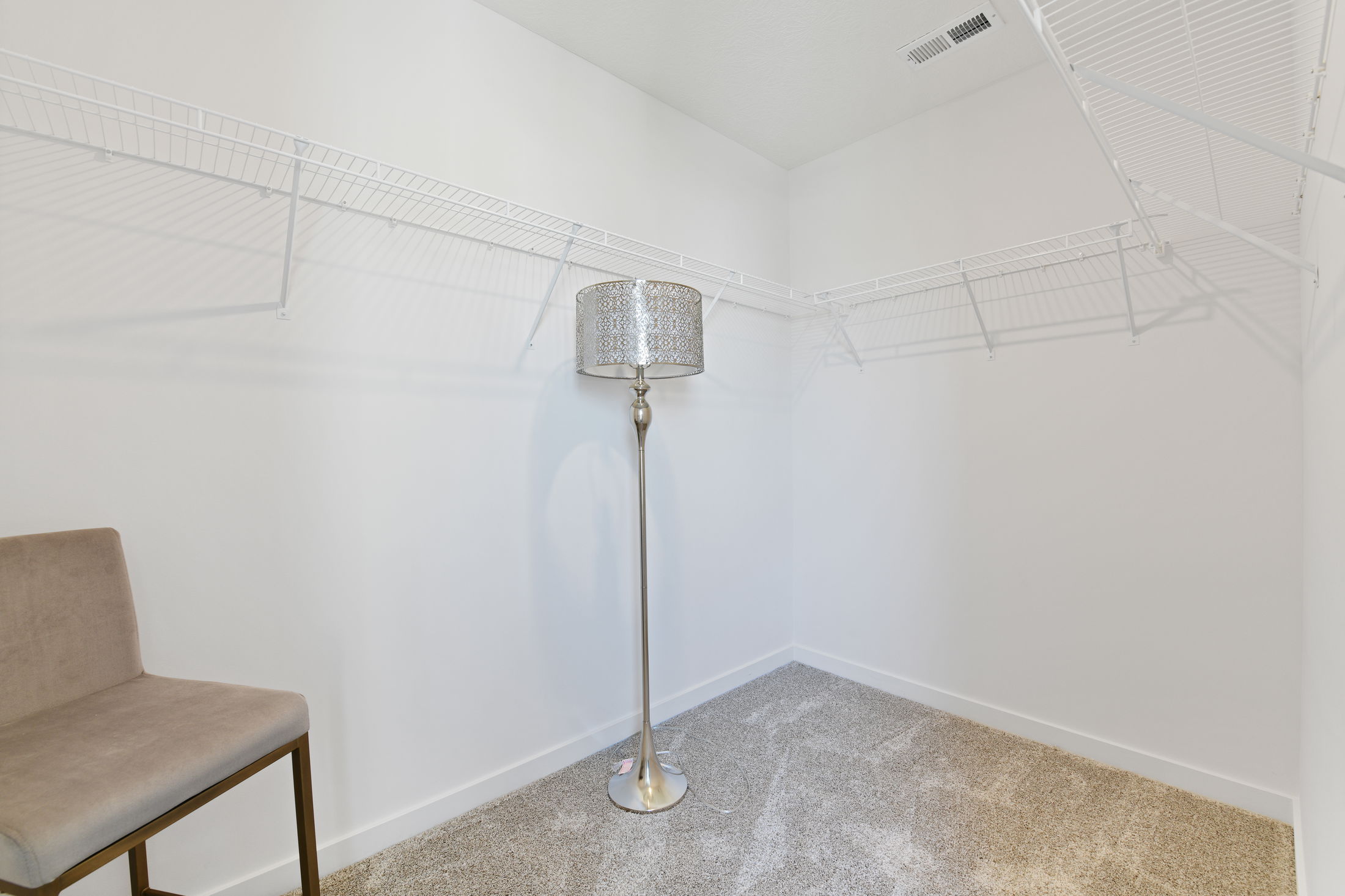 Empty walk-in closet with white wire shelving, carpeted floor, a modern floor lamp, and a beige chair.