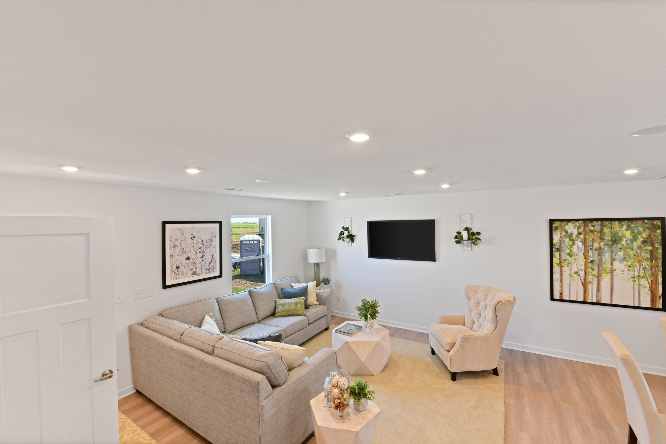 Bright, modern living room with a gray sectional, armchair, wall art, and a flat-screen TV on a white wall.