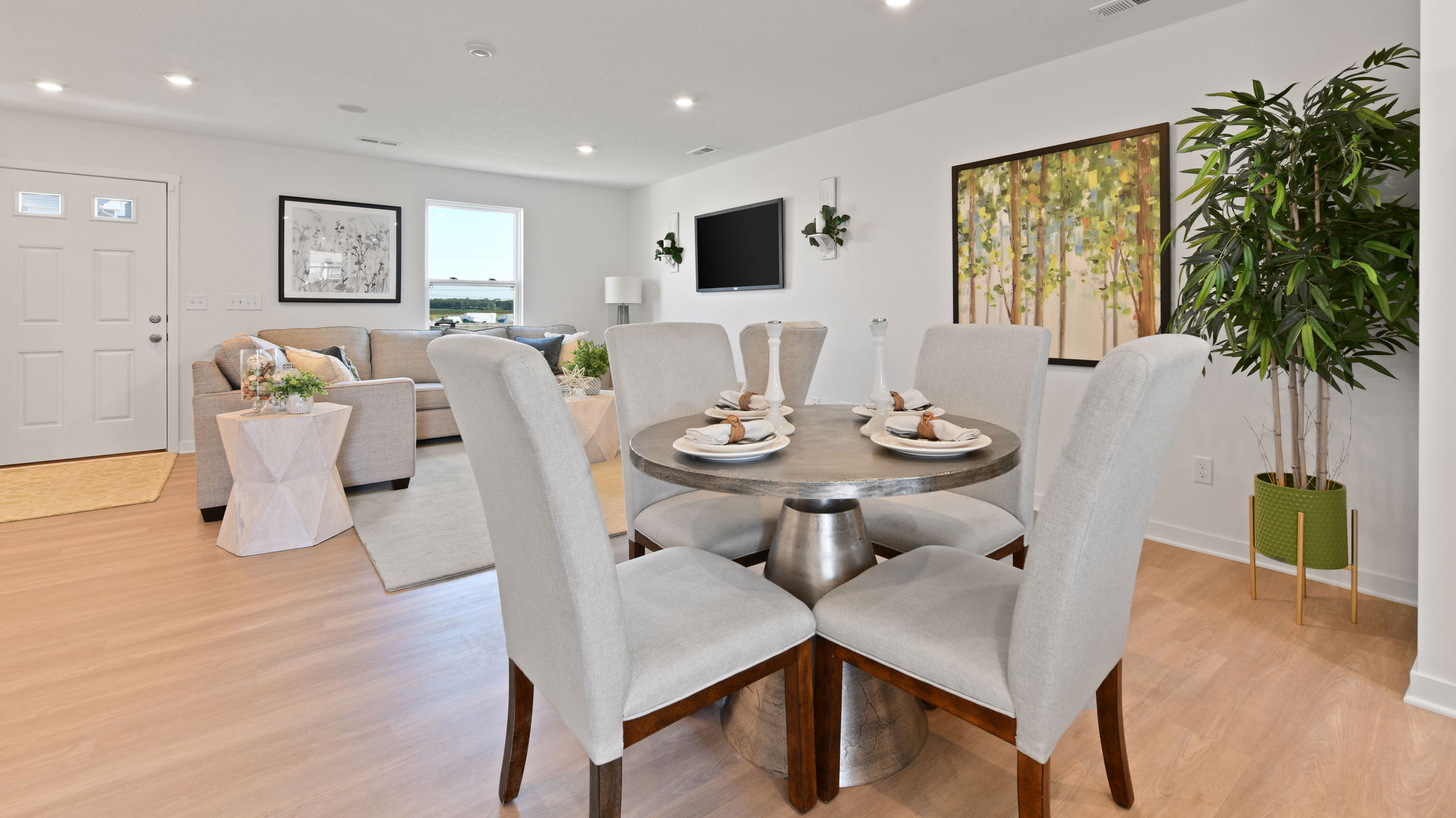 Modern open-plan living and dining area with neutral decor, featuring a round table set for four, light gray chairs, a cozy sofa, and vibrant wall art.