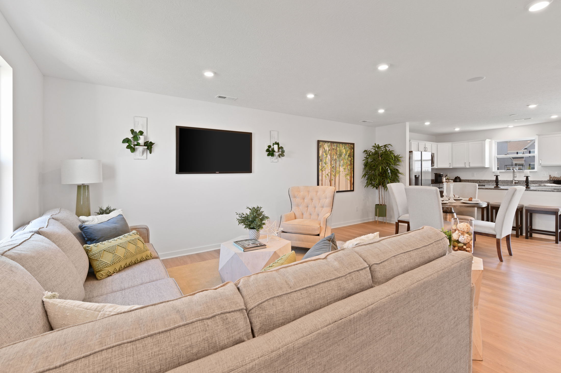 Modern open-plan living room with a beige sectional sofa, stylish armchair, wall-mounted TV, and kitchen area in the background.
