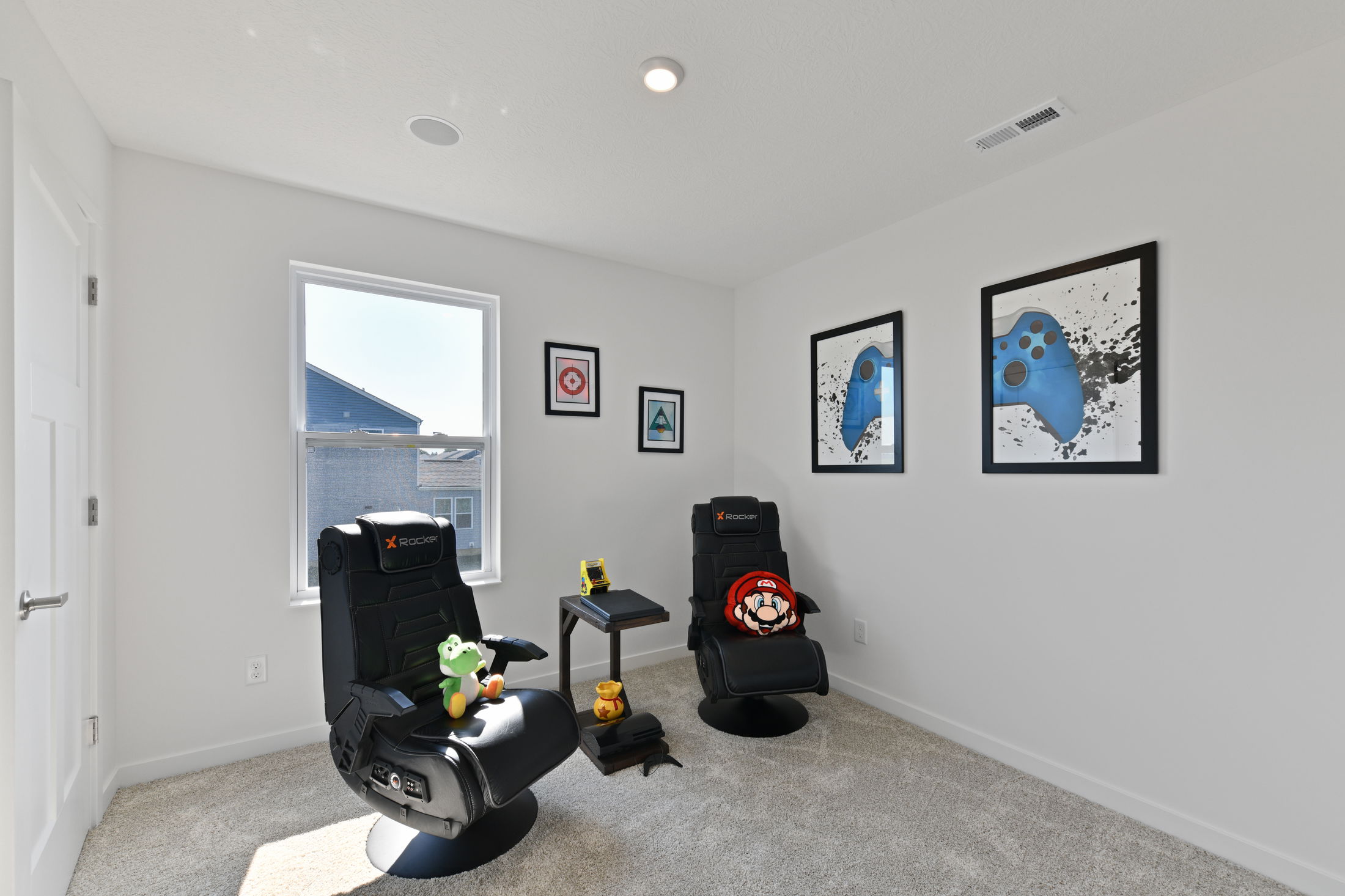 Gaming room setup featuring two black X Rocker chairs with character cushions, wall art, and a tabletop arcade machine by a window.