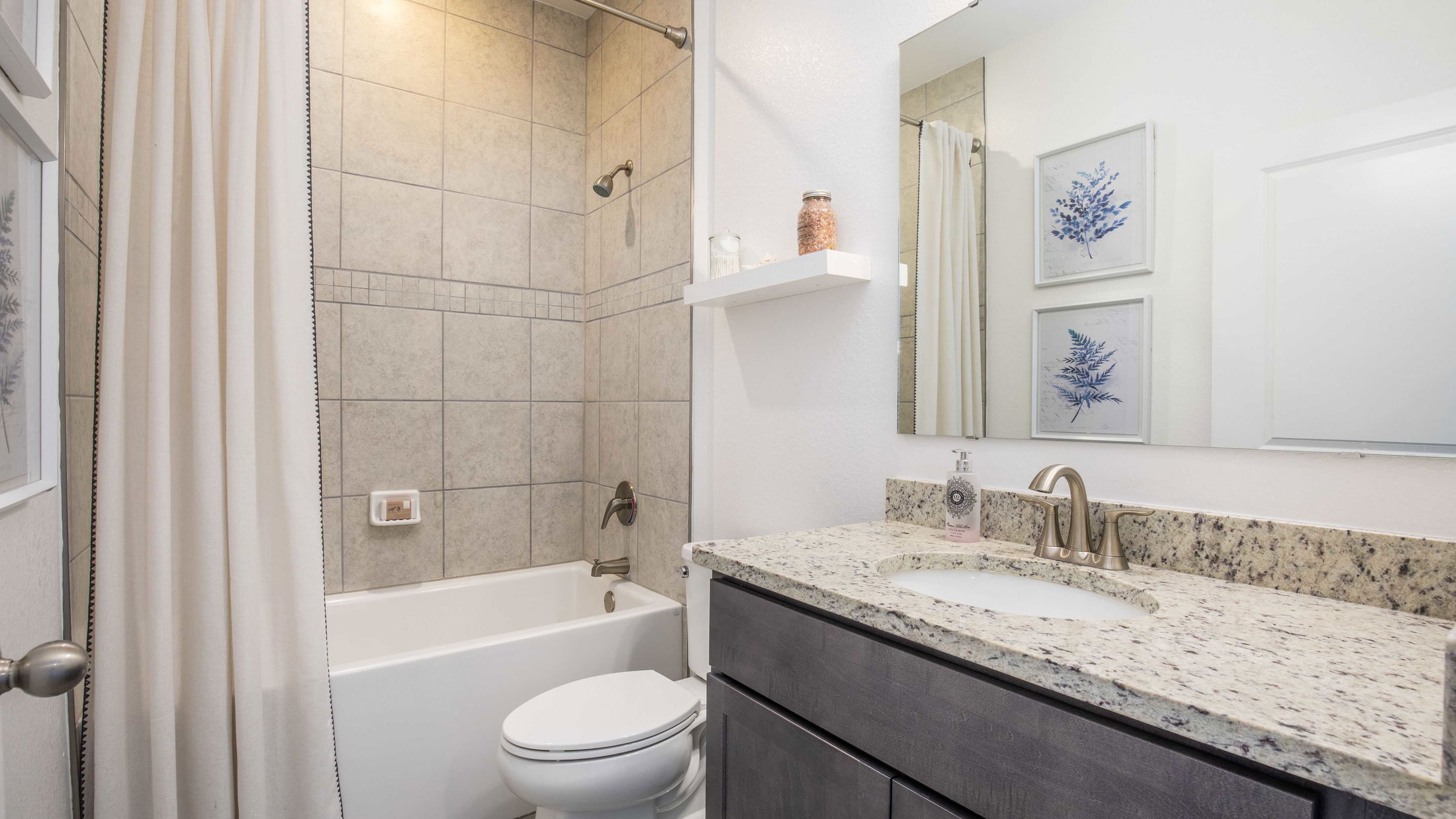 Modern bathroom with granite countertop, beige tiled bathtub, and decorative wall art.