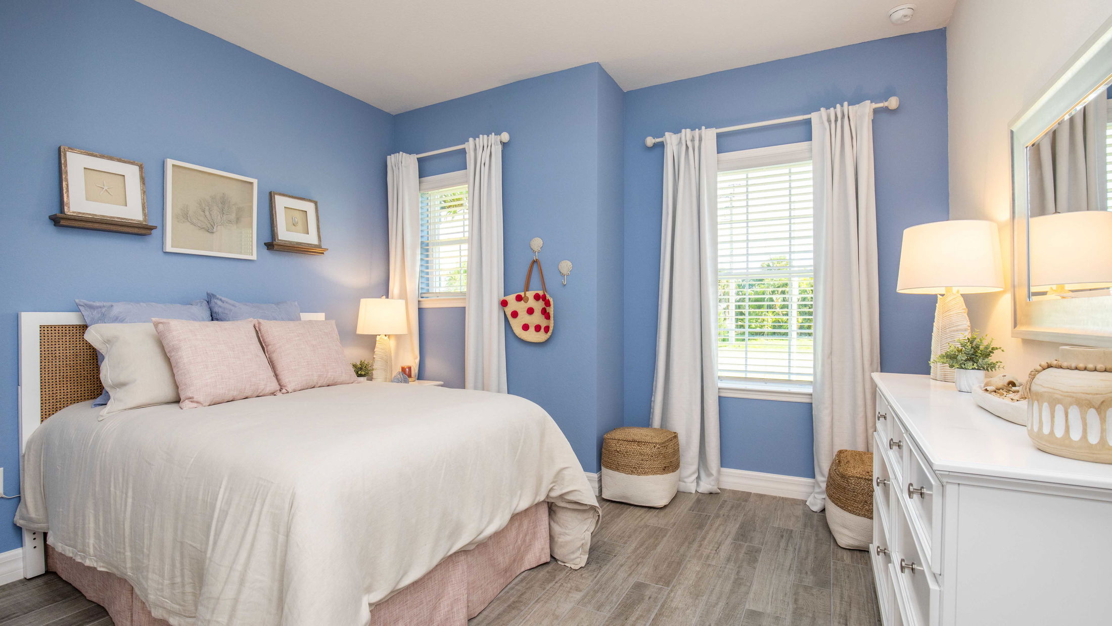 Cozy bedroom with a blue accent wall, beige bedding, wicker headboard, and bright natural light from two windows with white curtains.
