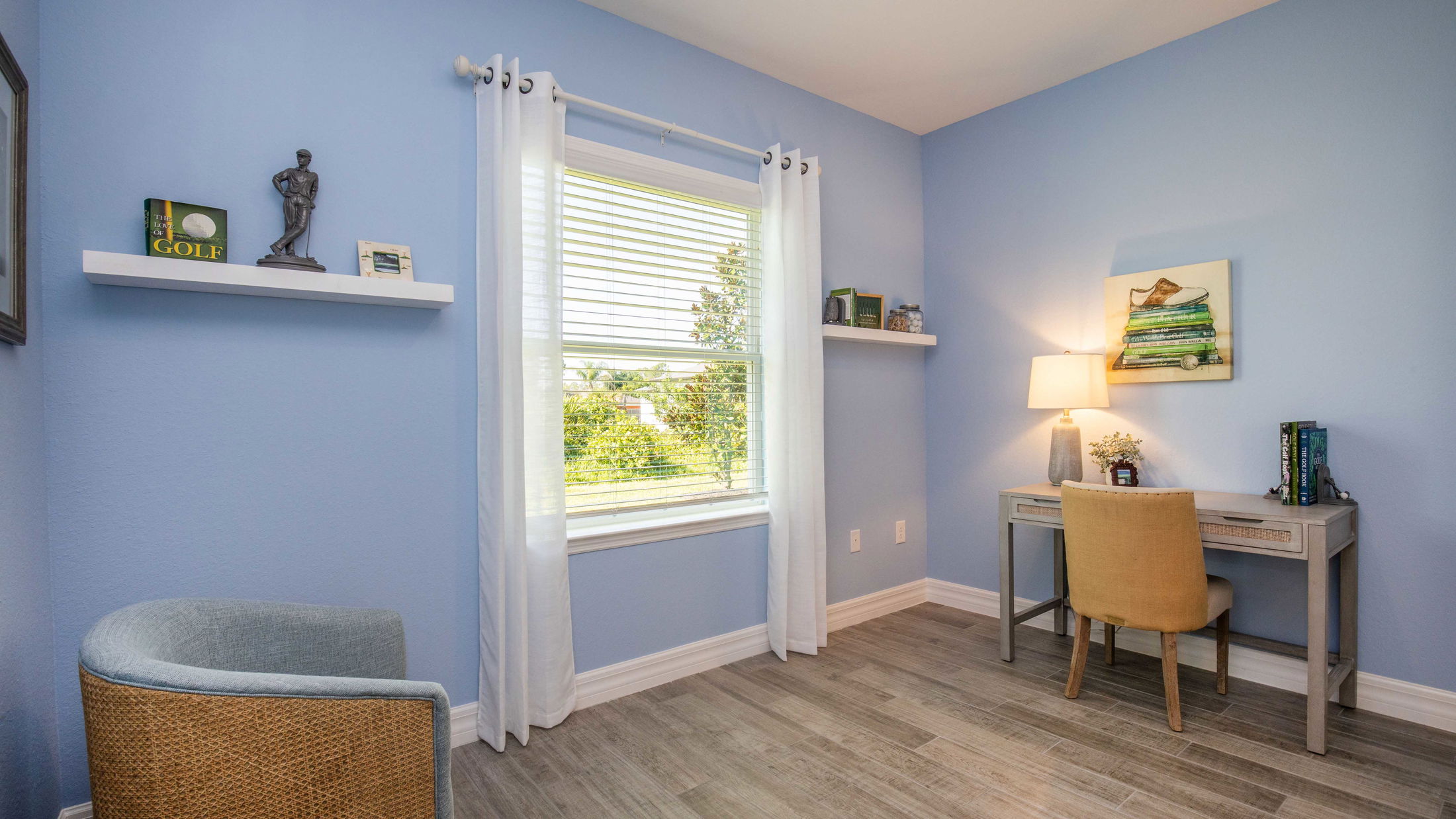Bright home office with wooden desk, chair, and decorative shelves, featuring a large window and blue walls.