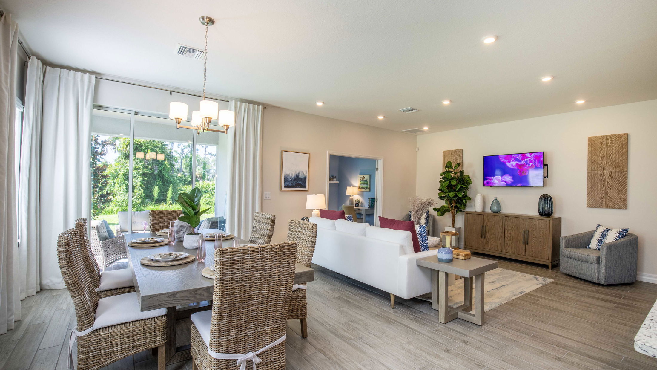 Modern open-concept living and dining room with wicker furniture, white and blue accents, and a garden view through large windows.