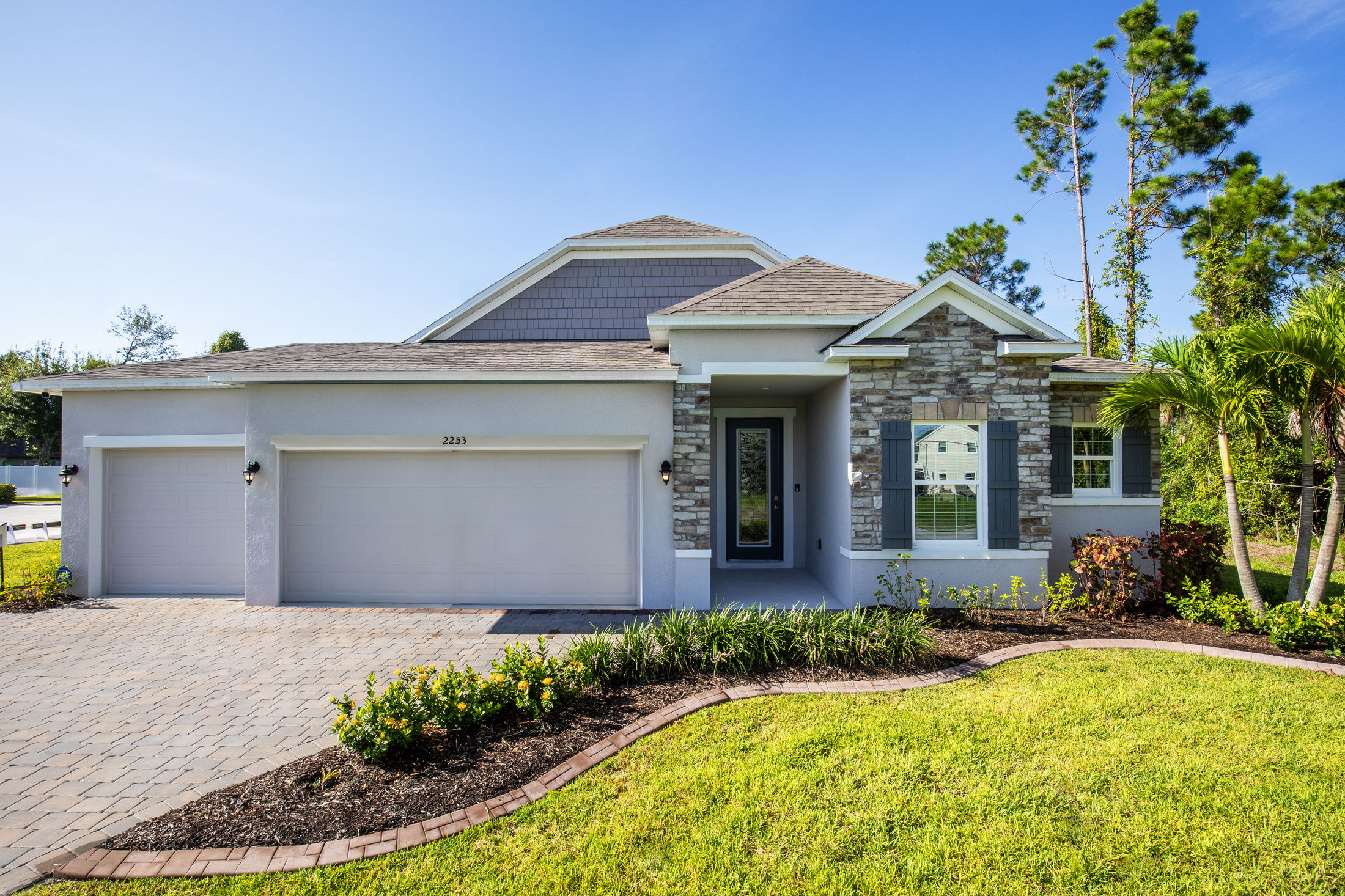 Modern single-story suburban home with a three-car garage, stone accents, and manicured lawn under a clear blue sky.