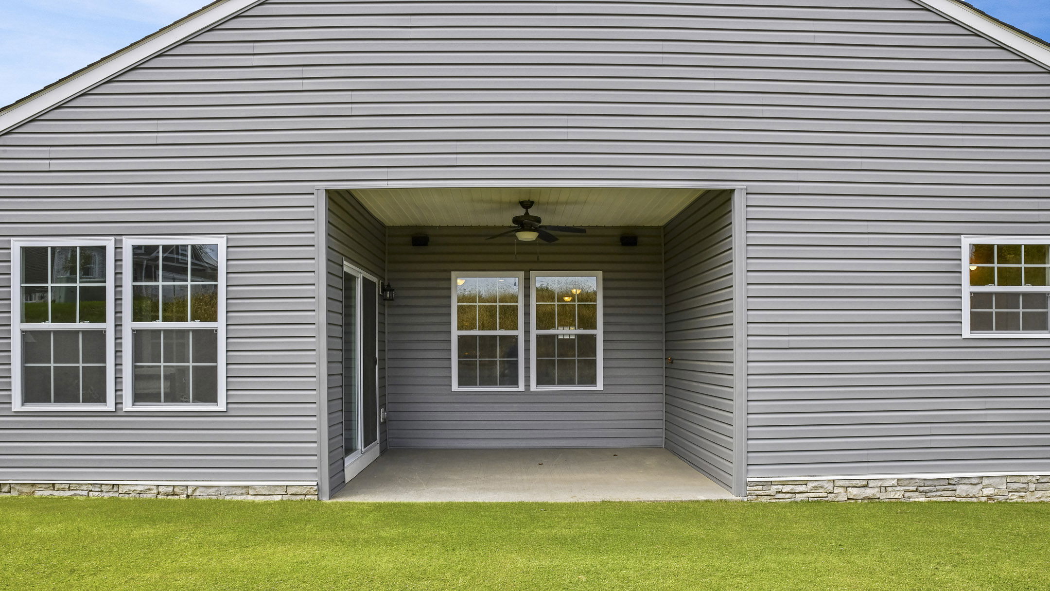 Gray house exterior featuring a covered patio with two windows, a ceiling fan, and a well-maintained lawn.
