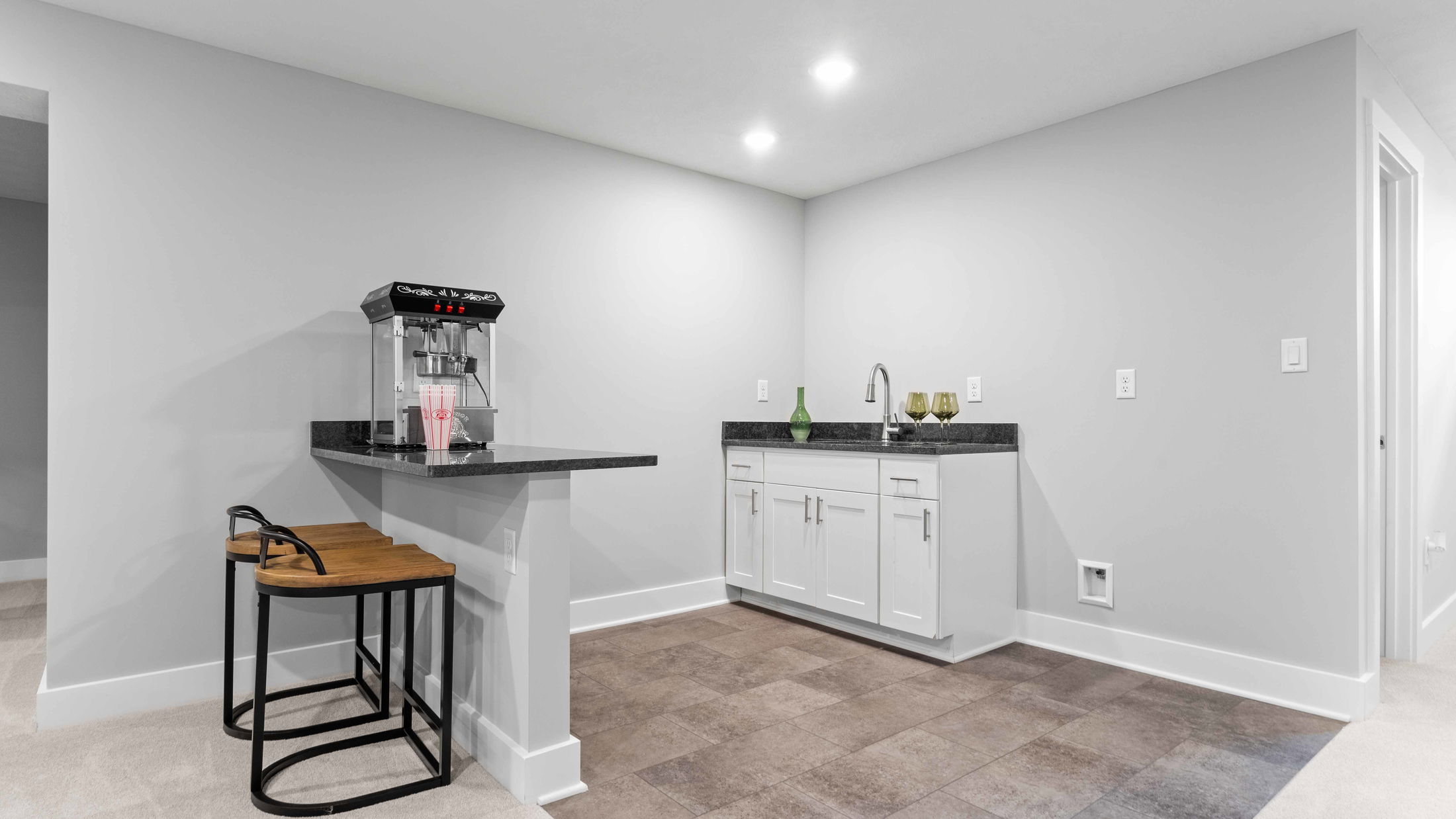 Modern basement kitchenette with popcorn machine, bar stools, and white cabinets.