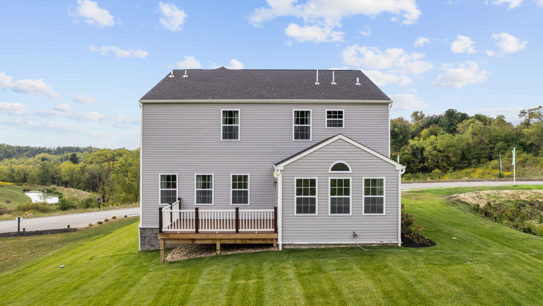 Two-story gray house with multiple windows and a small deck, surrounded by green grass and trees under a blue sky.