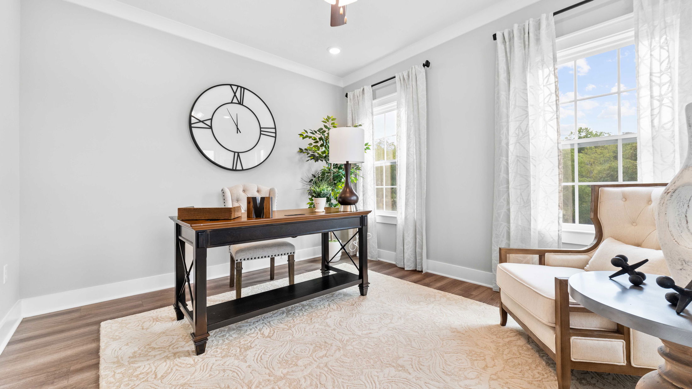 Stylishly decorated modern home office with a large wall clock, wooden desk, and cozy armchair by a window with natural light.