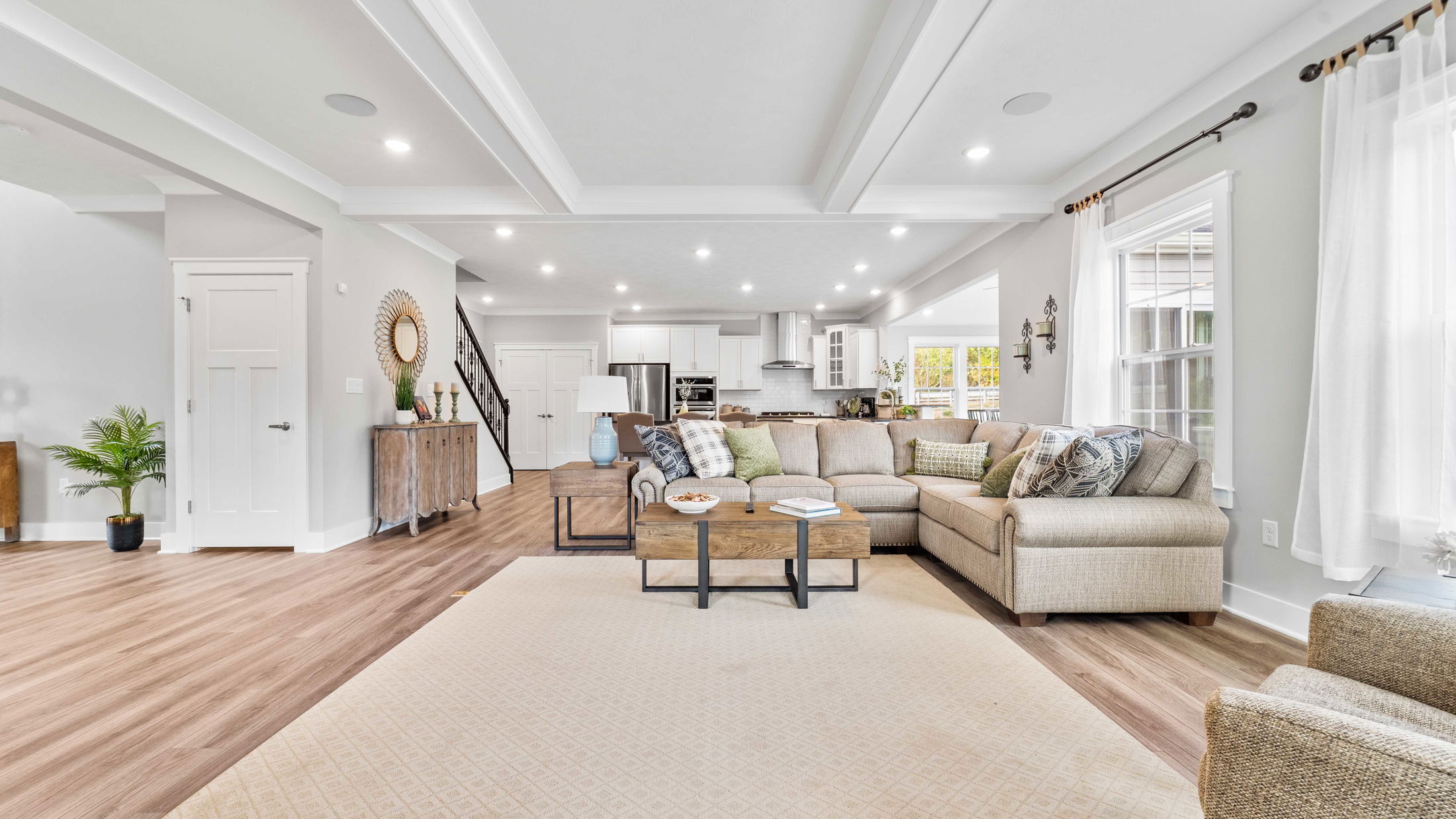 Modern open-concept living room with beige sectional sofa, wooden coffee table, and kitchen in the background, featuring elegant decor and abundant natural light.