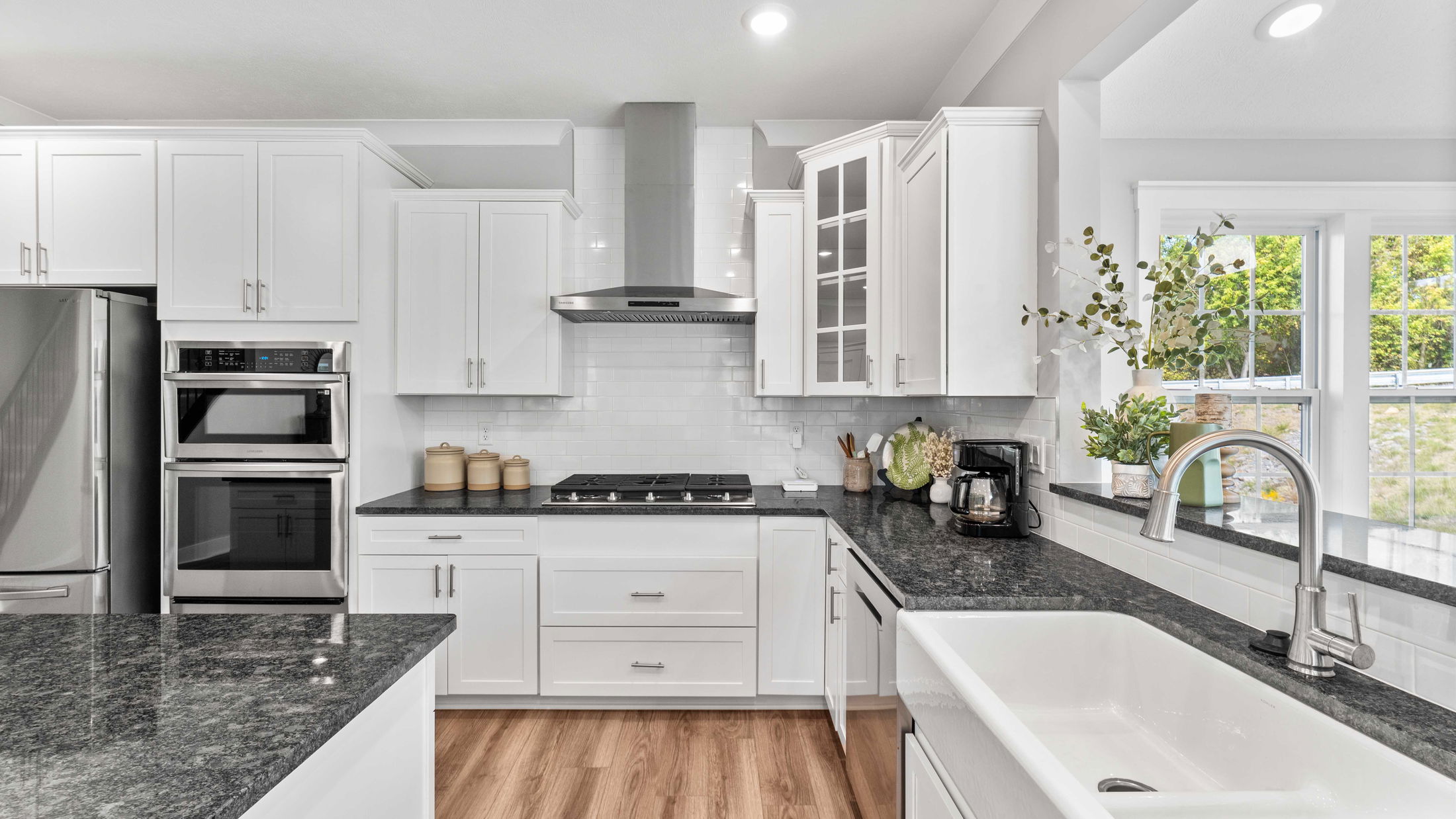 Modern white kitchen with stainless steel appliances, black granite countertops, and a farmhouse sink near sunny windows.