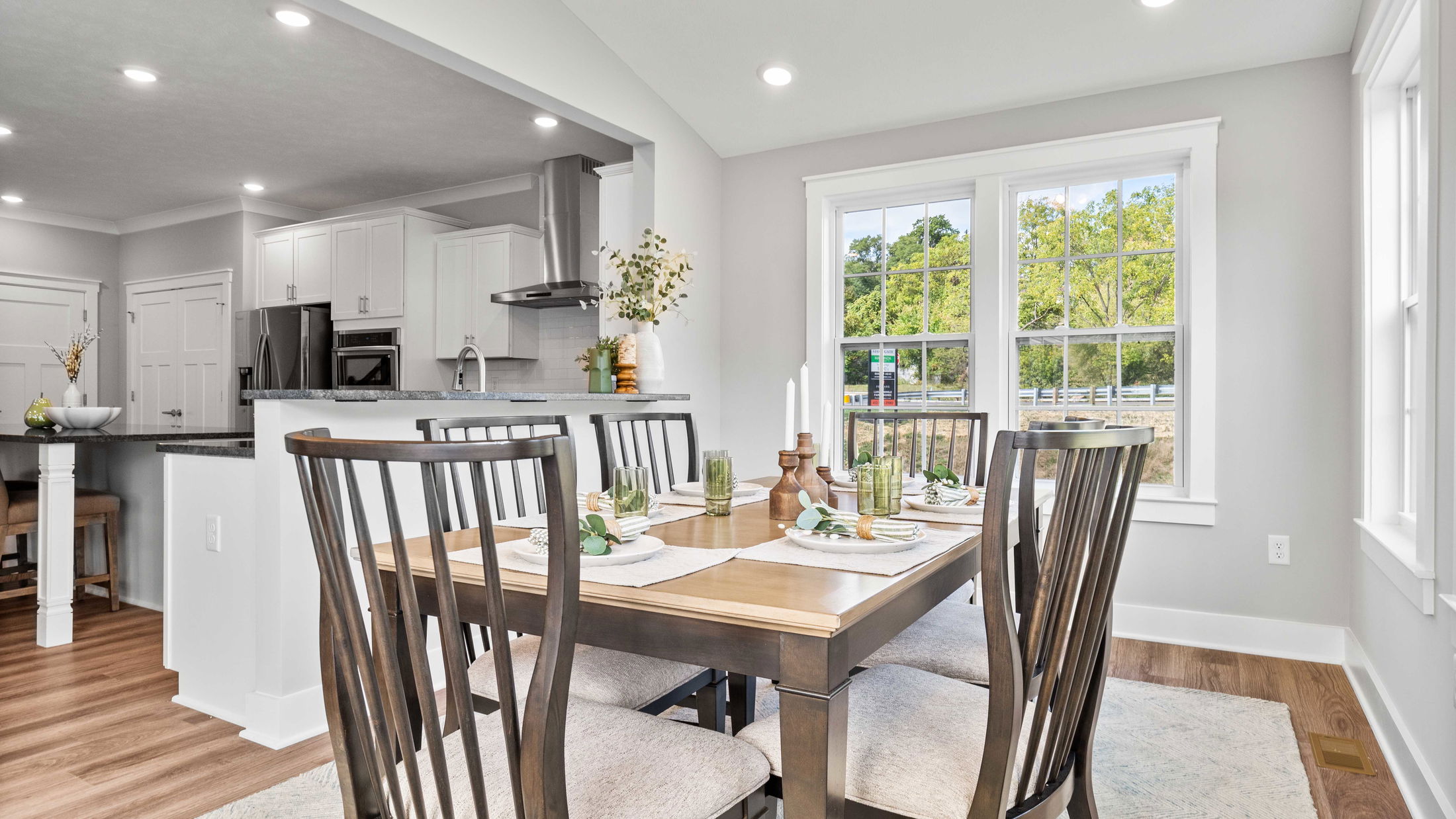 Modern dining area with wooden table and chairs, open to a sleek kitchen with stainless steel appliances and large windows offering a view of greenery.