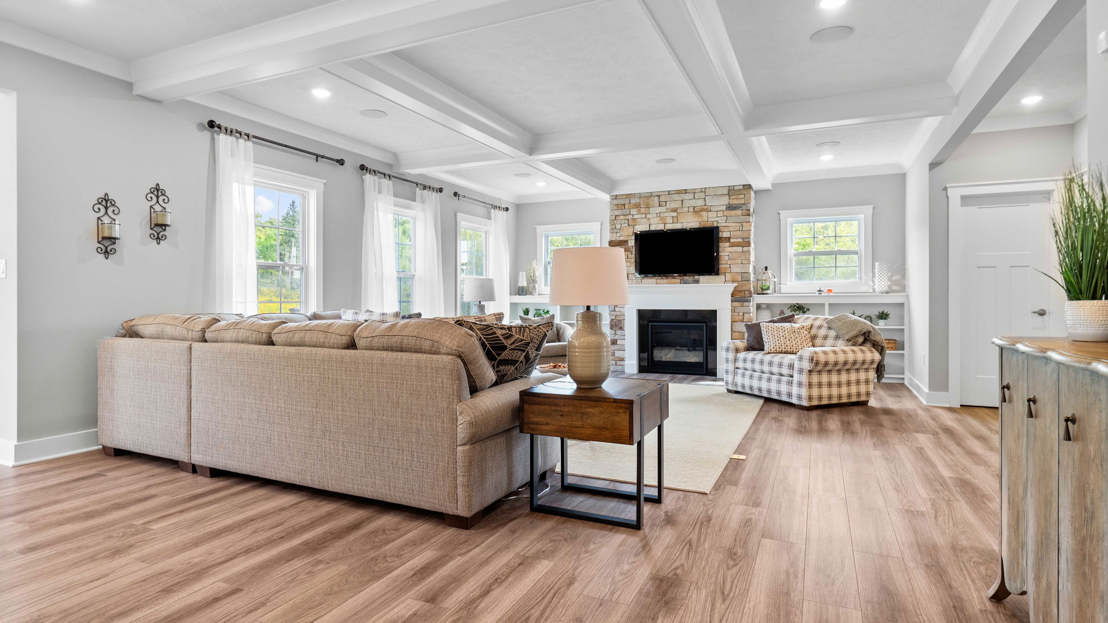 Modern living room interior with a large beige sectional sofa, wood flooring, and a stone fireplace under a coffered ceiling.