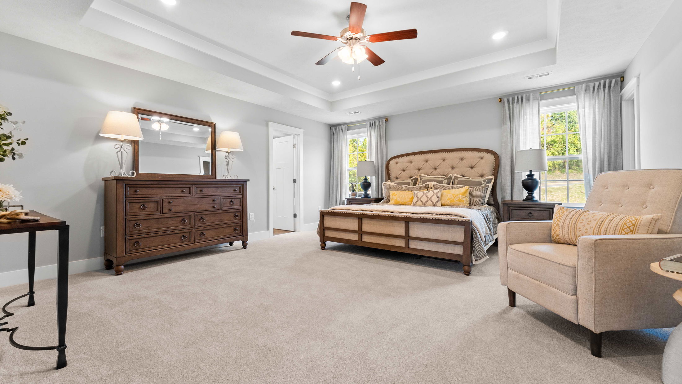 Spacious bedroom with elegant decor, featuring a large bed, wooden dresser, and cozy armchair under a ceiling fan.