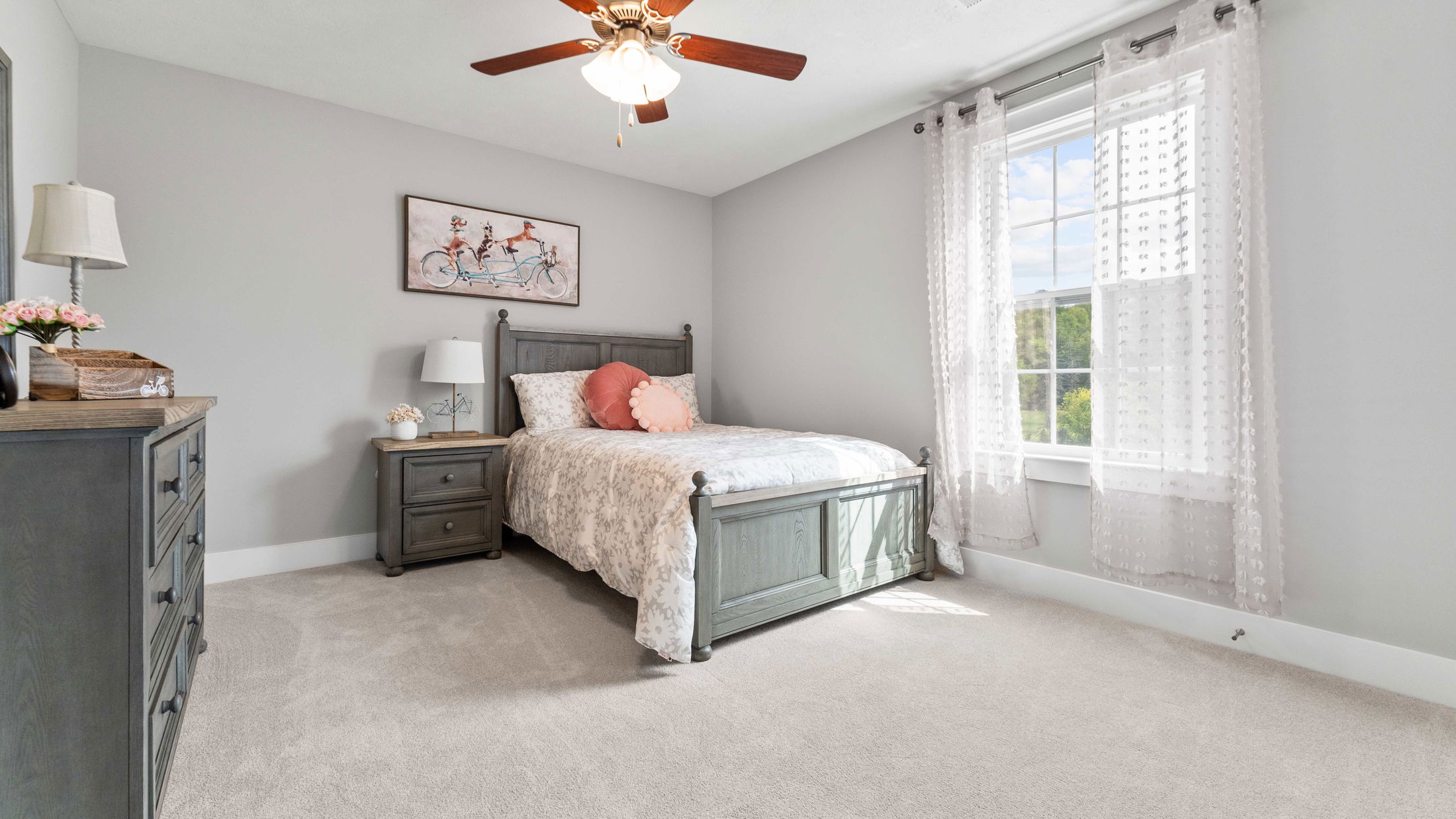 Bright and cozy bedroom with stylish gray furniture, floral bedding, and natural light streaming through a window with sheer curtains.