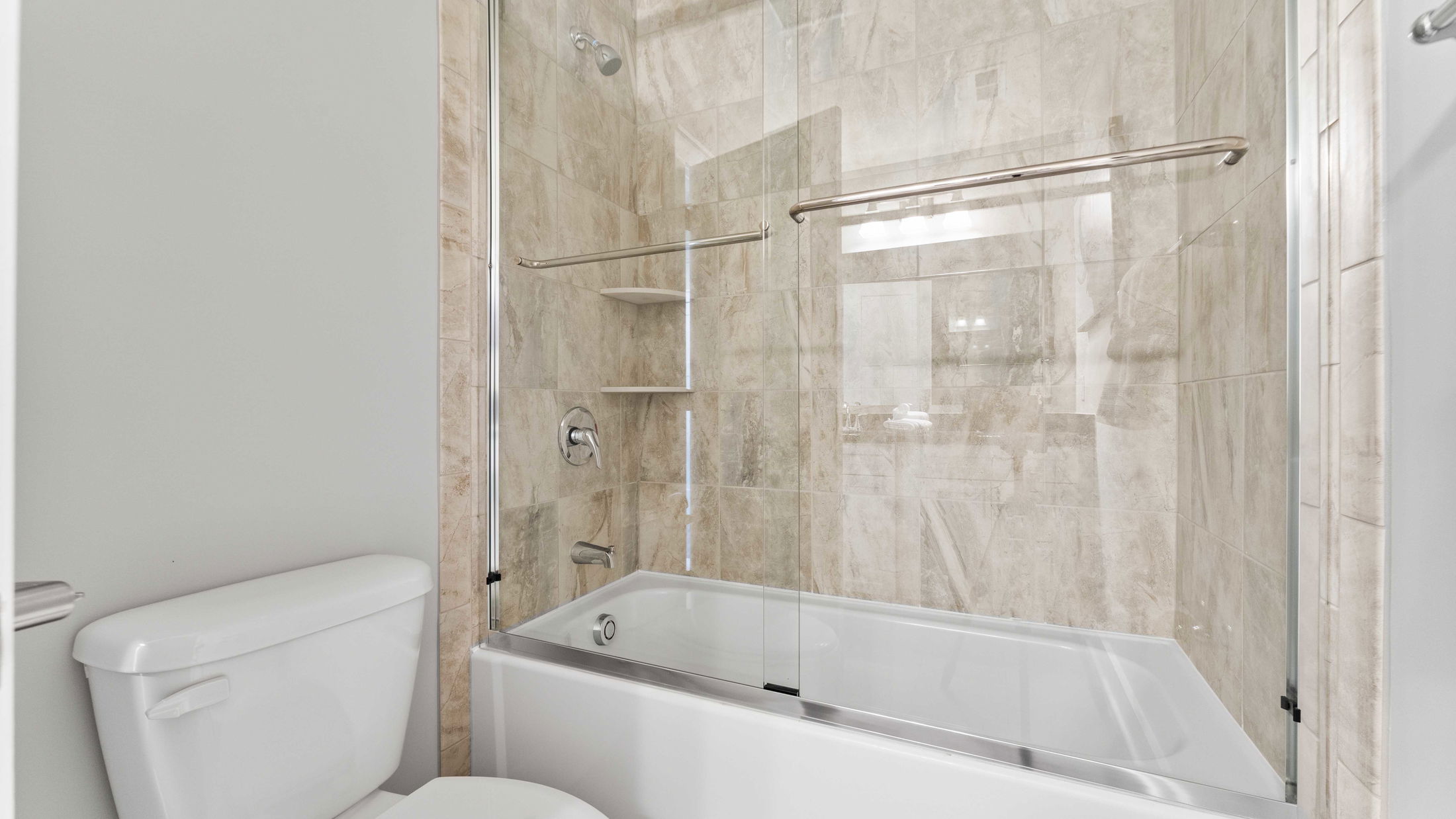 Modern bathroom with a tiled bathtub and glass shower door, featuring chrome fixtures and a white toilet.