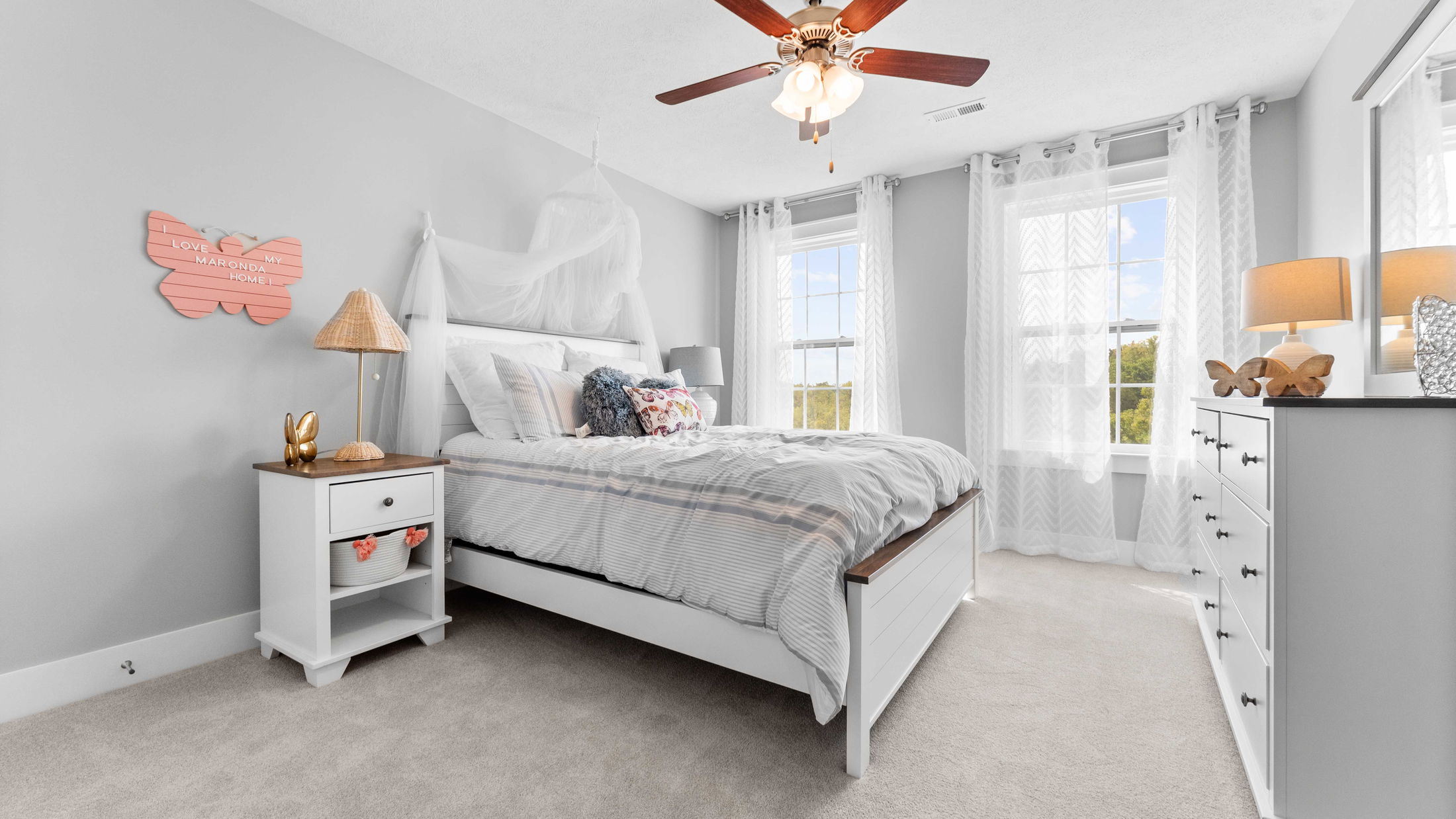 Modern, airy bedroom with a white bed, ceiling fan, and natural light streaming through sheer curtains.