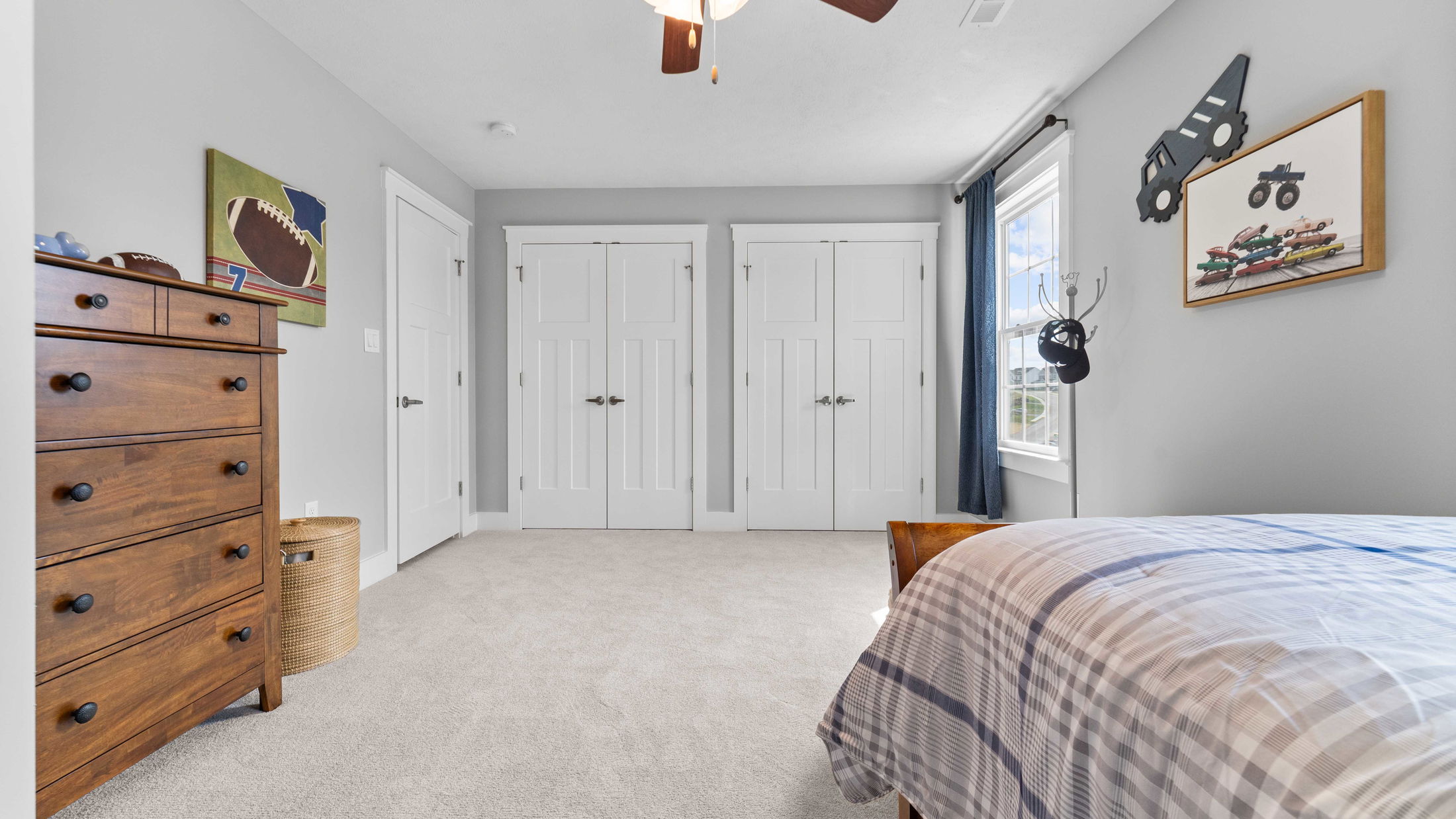 Cozy gray kids' bedroom with a wooden dresser, football-themed decor, and plush carpet, featuring double closets and a plaid bedspread.