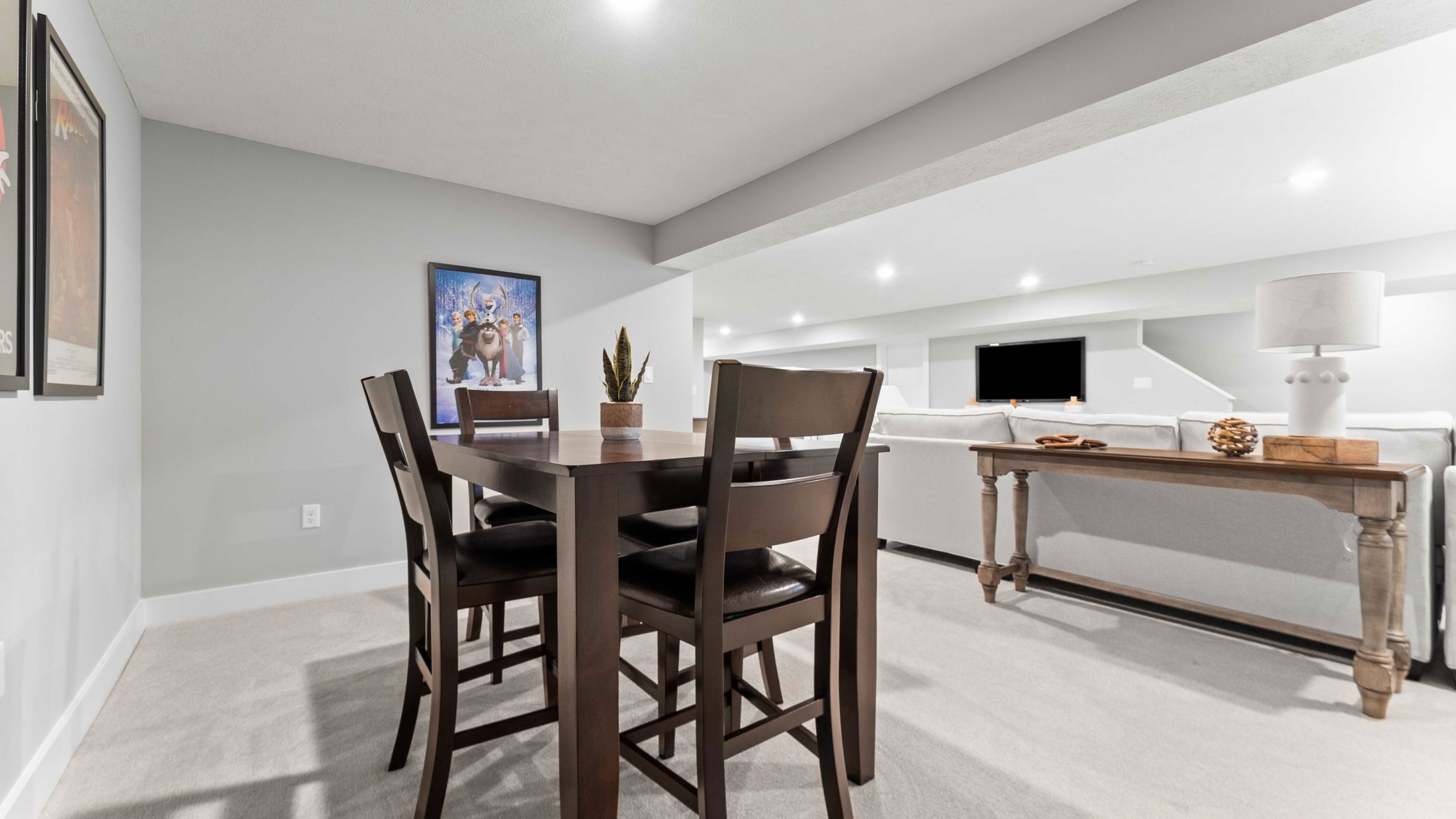 A modern, well-lit basement recreation room with a wooden dining table set, neutral-colored sofas, a long console table, and framed wall art.